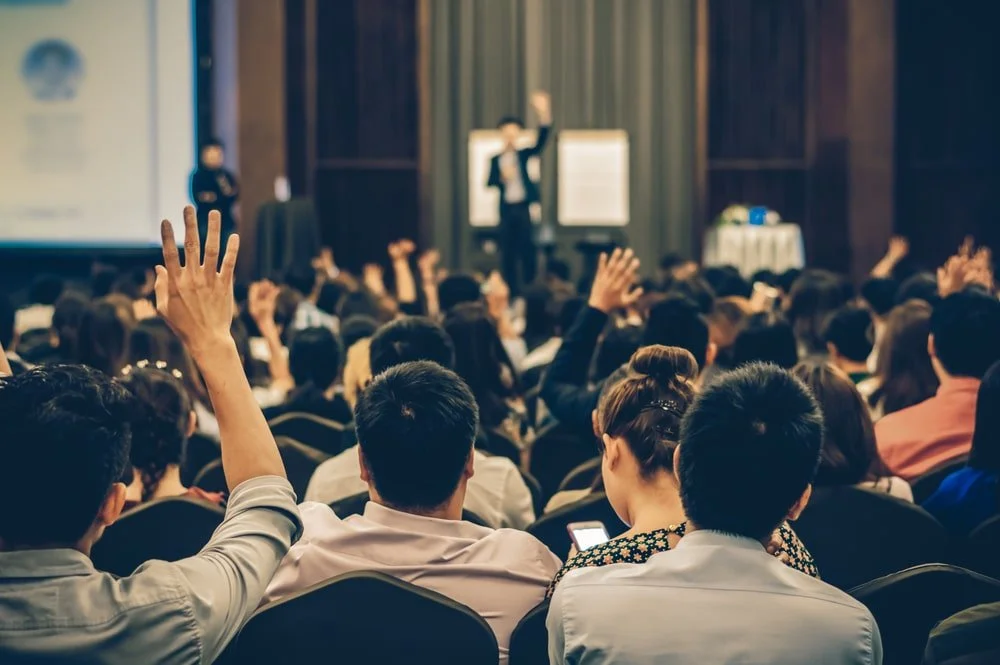 A large group of people in a conference hall, some raising their hands, attending a presentation with a speaker at the front. giving a cpd certified course