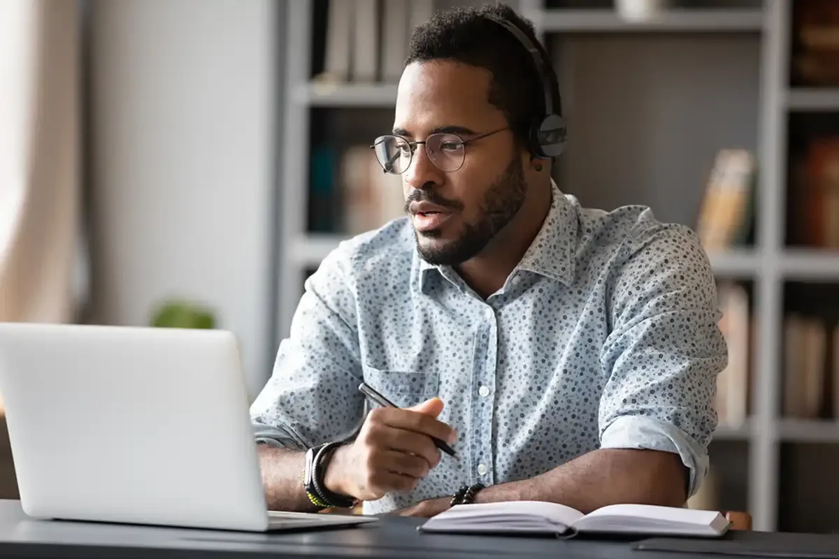 Man wearing glasses and headphones studying or working at a desk with a laptop and open CPD book.