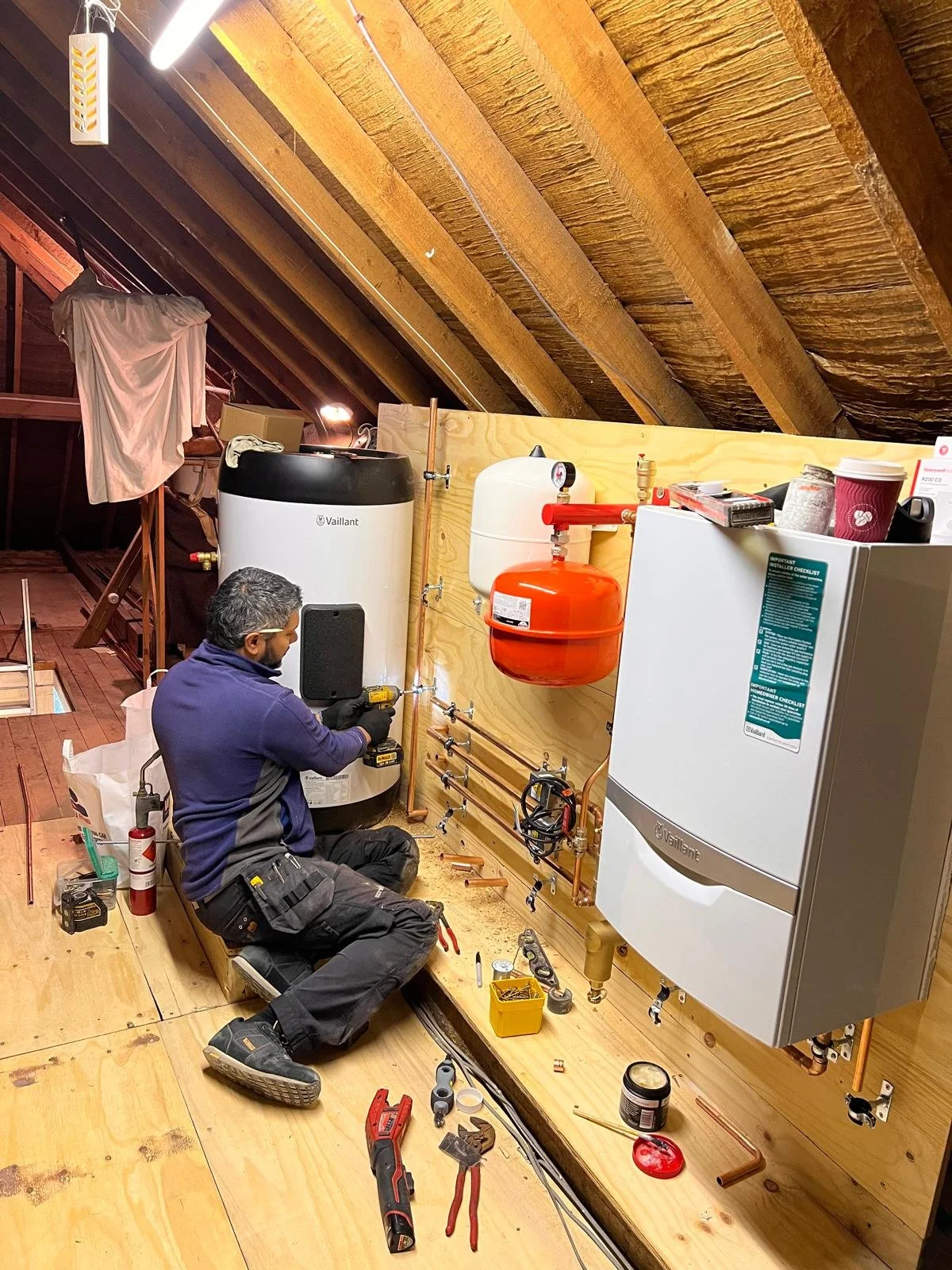 A technician installing or repairing a water heating system in an attic with wooden flooring and sloped ceiling. Various tools are scattered around, and there are large white and orange water tanks and pipes.