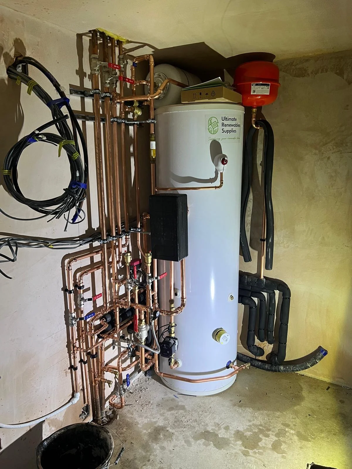 A water heater and plumbing system in a utility room with copper pipes, a black box, and insulated pipes, set against unfinished concrete walls.