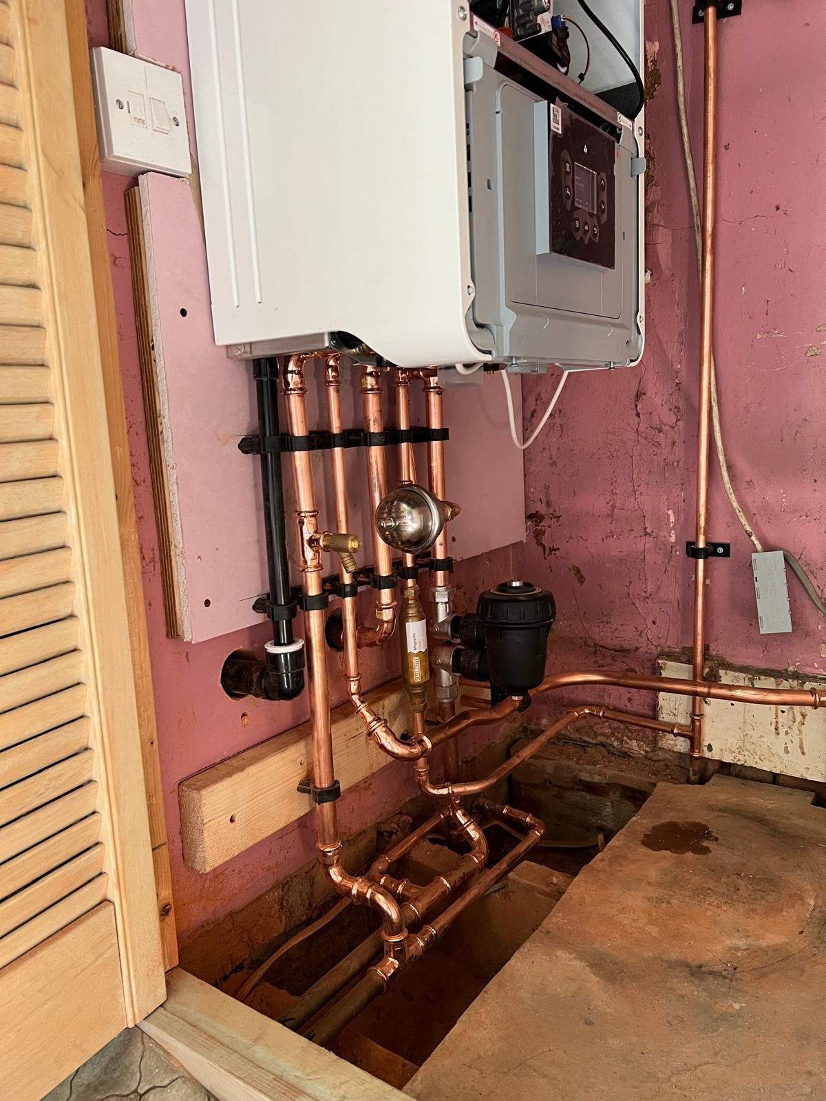 A plumbing and heating system with copper pipes, a boiler, and a pressure tank installed in a basement with a pink wall and exposed flooring.