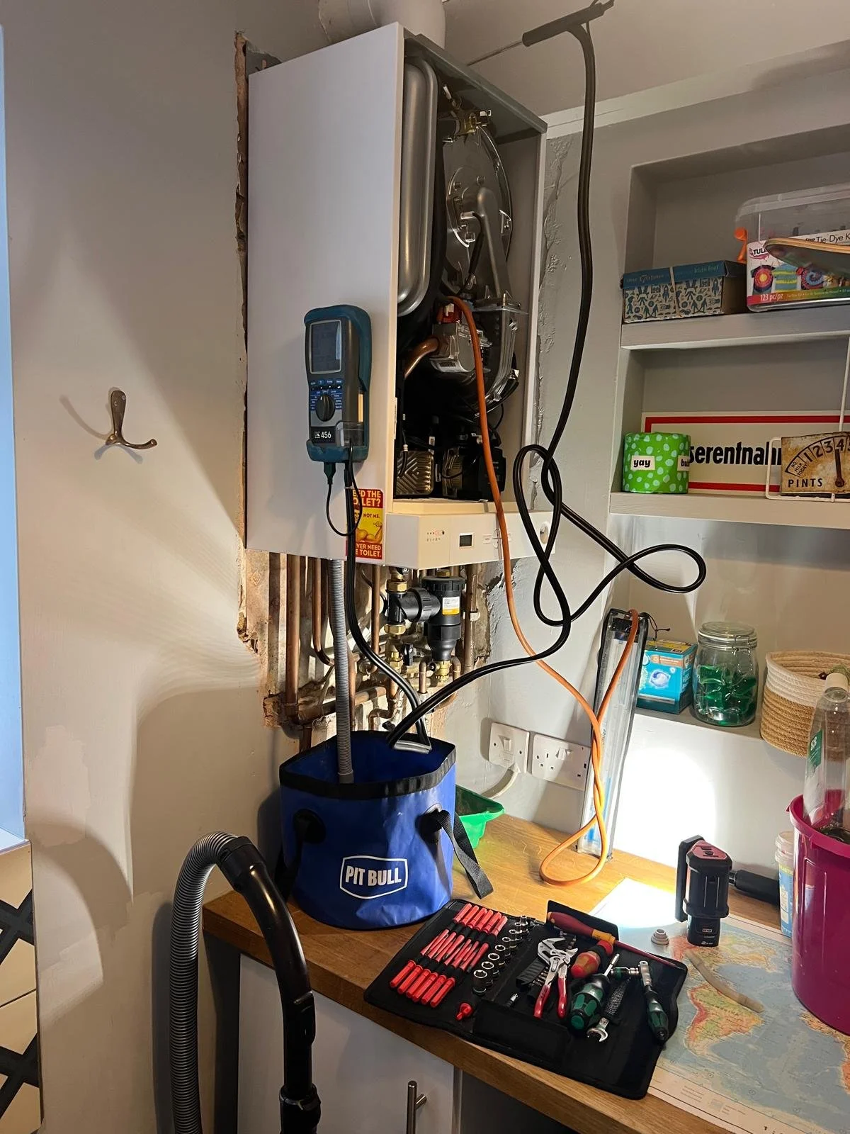 An electrical or plumbing device installed inside a cutout in the wall, with tools and a blue Pit Bull tool bag on the countertop below, and various tools and supplies around.
