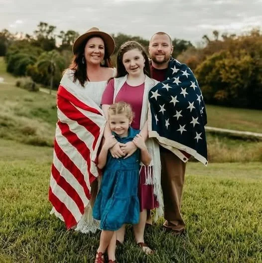 Four people standing outdoors on grass with trees and an overcast sky in background. Two women and two children, one girl and one boy, are smiling. The woman on the left is wearing a hat and an American flag shawl, the girl in front is dressed in a blue dress, the girl in the middle has a pink top and red dress, and the man on the right is wrapped in an American flag.