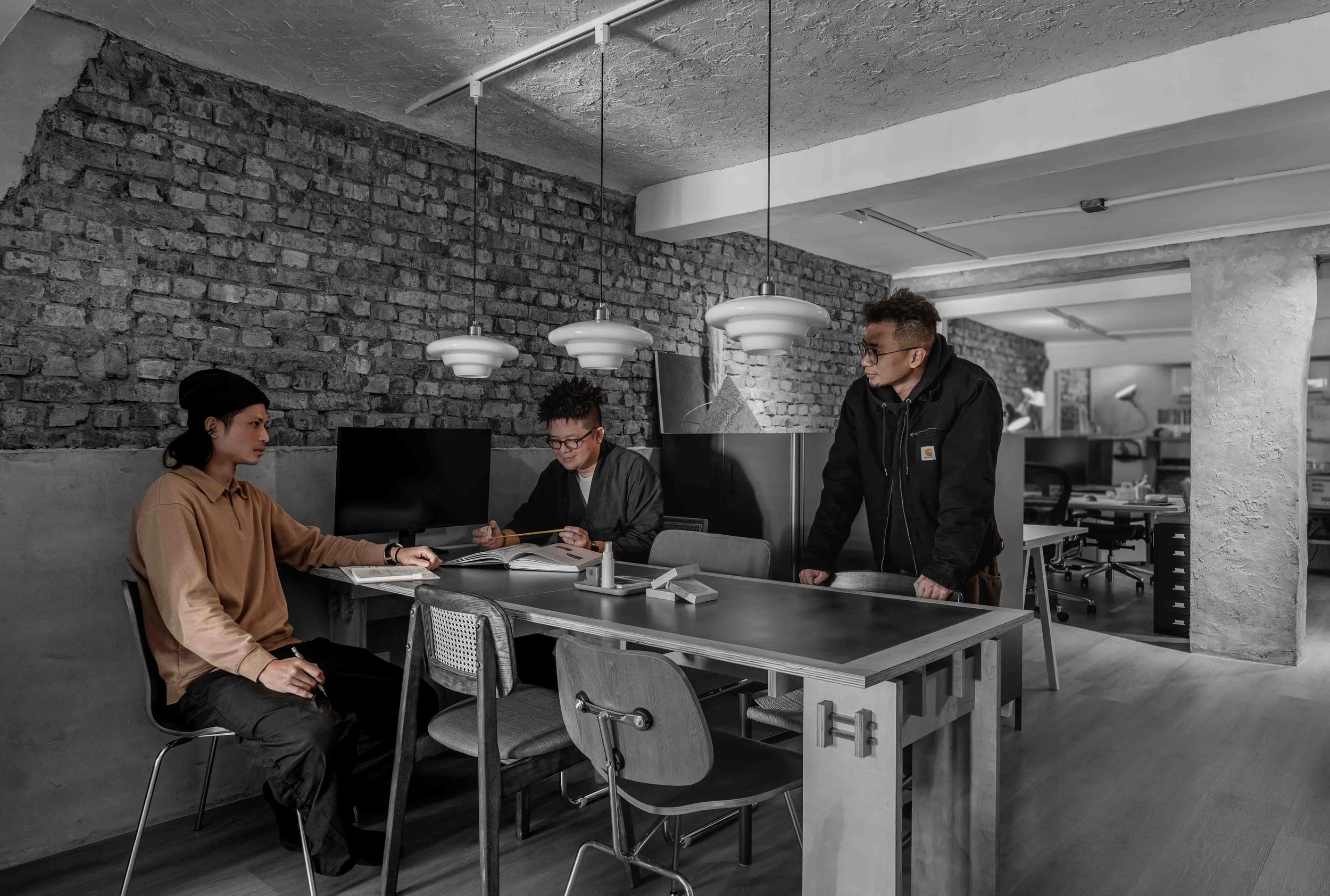 Three people in an mid century modern office meeting room with a brick wall and pendant lights. Two sit at a table, one standing.
