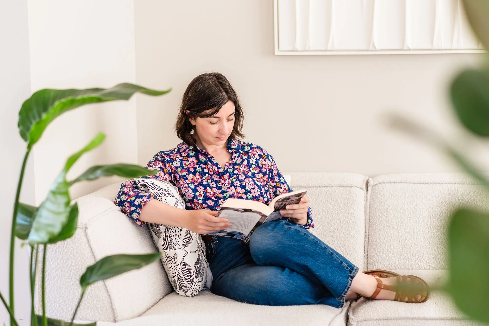 A neurodivergent woman with shoulder-length brown hair in a floral blouse sitting on a light-colored sofa with her legs tucked up under her, reading a book. The room is bright with white walls and green plants in the foreground.