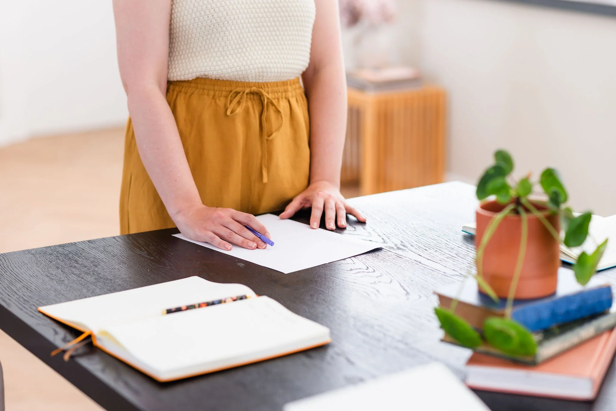 A neurodivergent woman stands at the head of a table that is set for a group meeting, with a stack of books, a potted plant, and an open notebook and pen. Her face is not visible and her hands rest on the table with confidence.