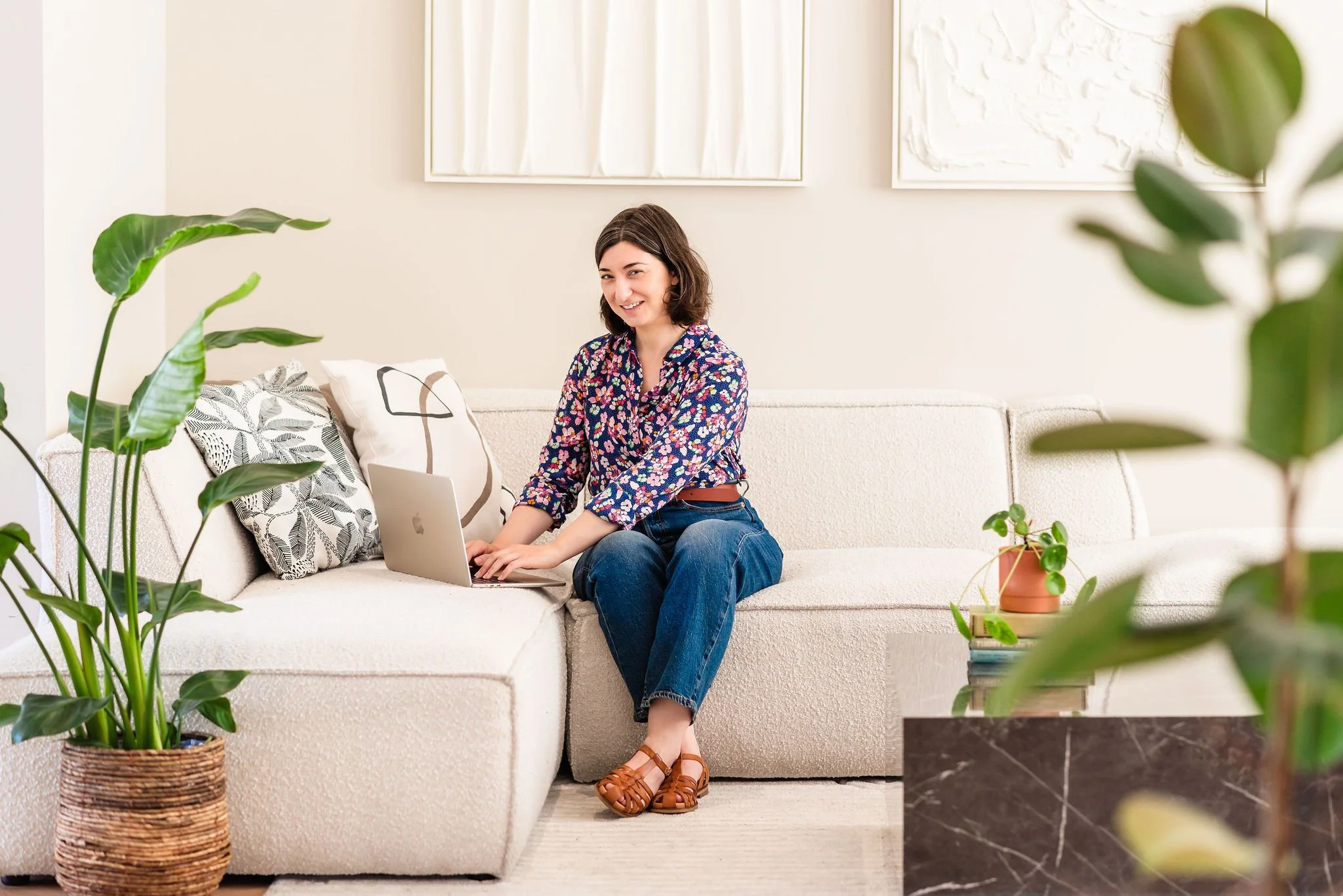 neurodivergent woman wearing jeans and a colorful floral blouse works on a laptop while sitting on a white sofa in a modern, well-lit living room. She pauses in her work to smile at the camera.