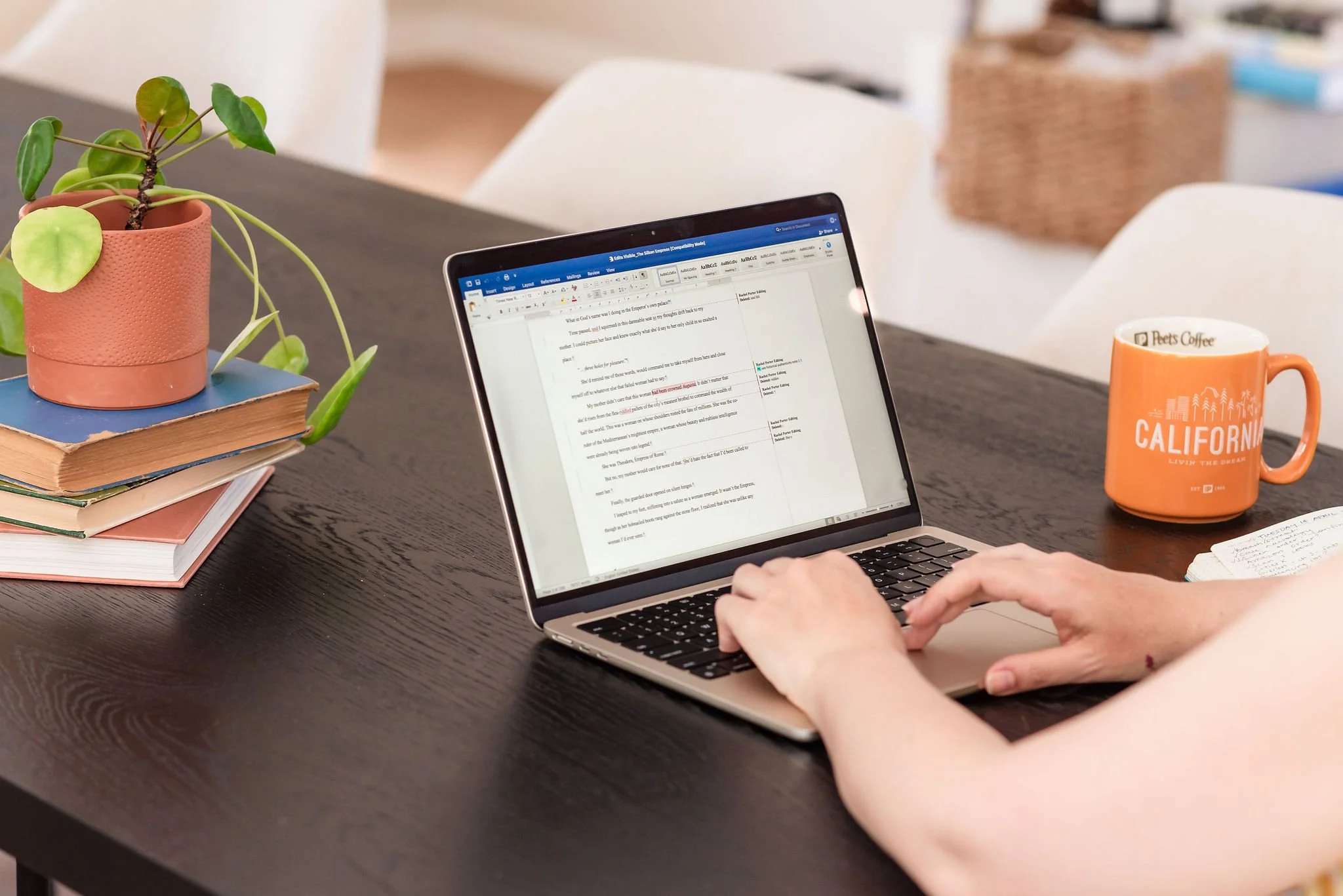A woman edits a document on a laptop at a desk. Only her hands are visible; a stack of books, a potted plant, and an orange CALIFORNIA coffee mug are just out of shot beside her.