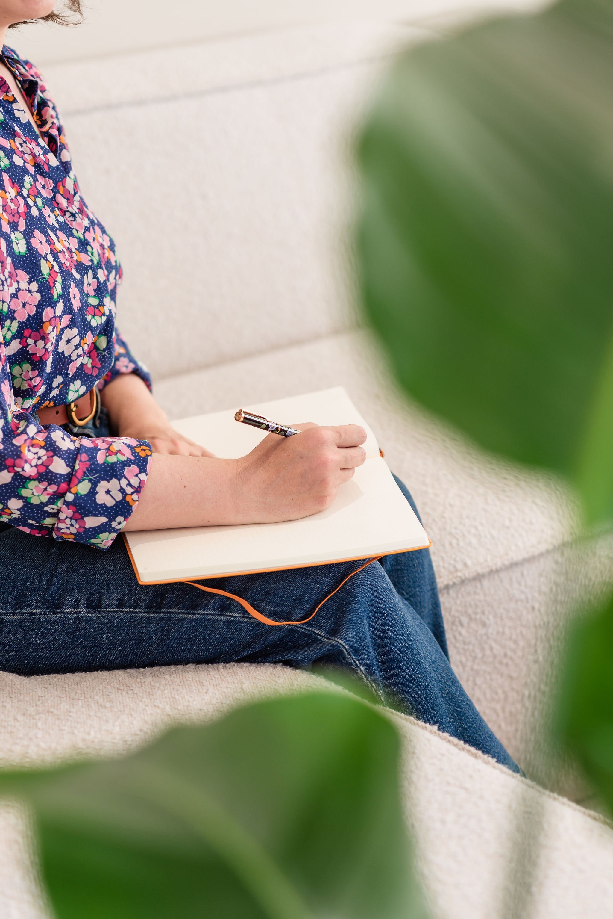 Woman whose face is out of frame, wearing a floral shirt sitting on a beige couch, writing in a notepad with a pen.
