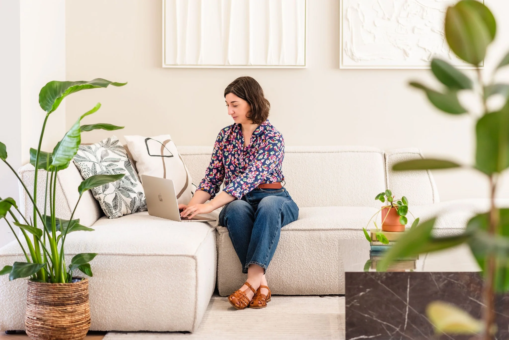 A neurodivergent woman with shoulder-length brown hair sitting on a white sofa working on a laptop, surrounded by large potted plants in a bright, modern living room.