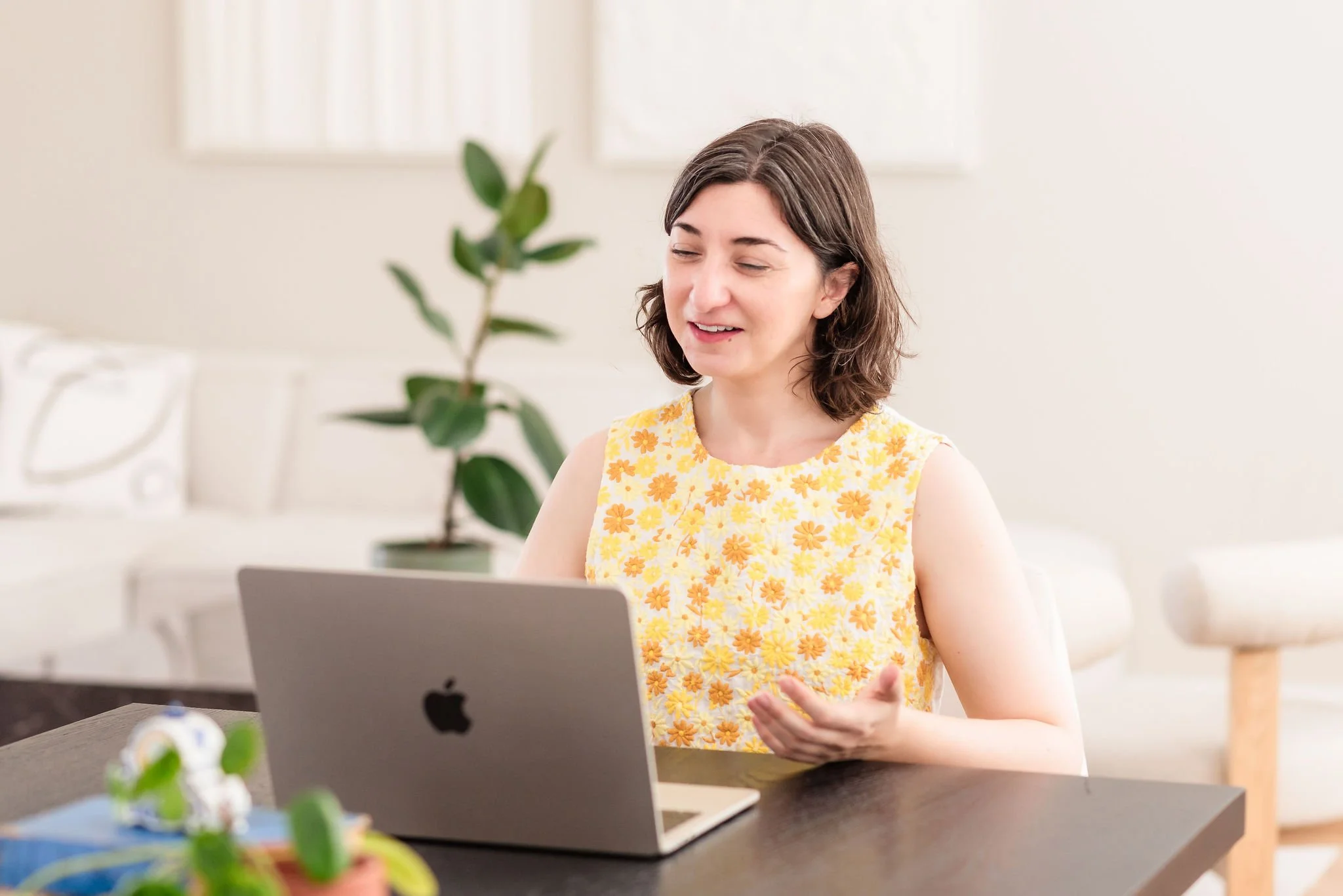 A neurodivergent woman with shoulder-length brown hair wearing a yellow floral top sitting at a desk, smiling and talking into a laptop in front of her, with a potted plant in the background.