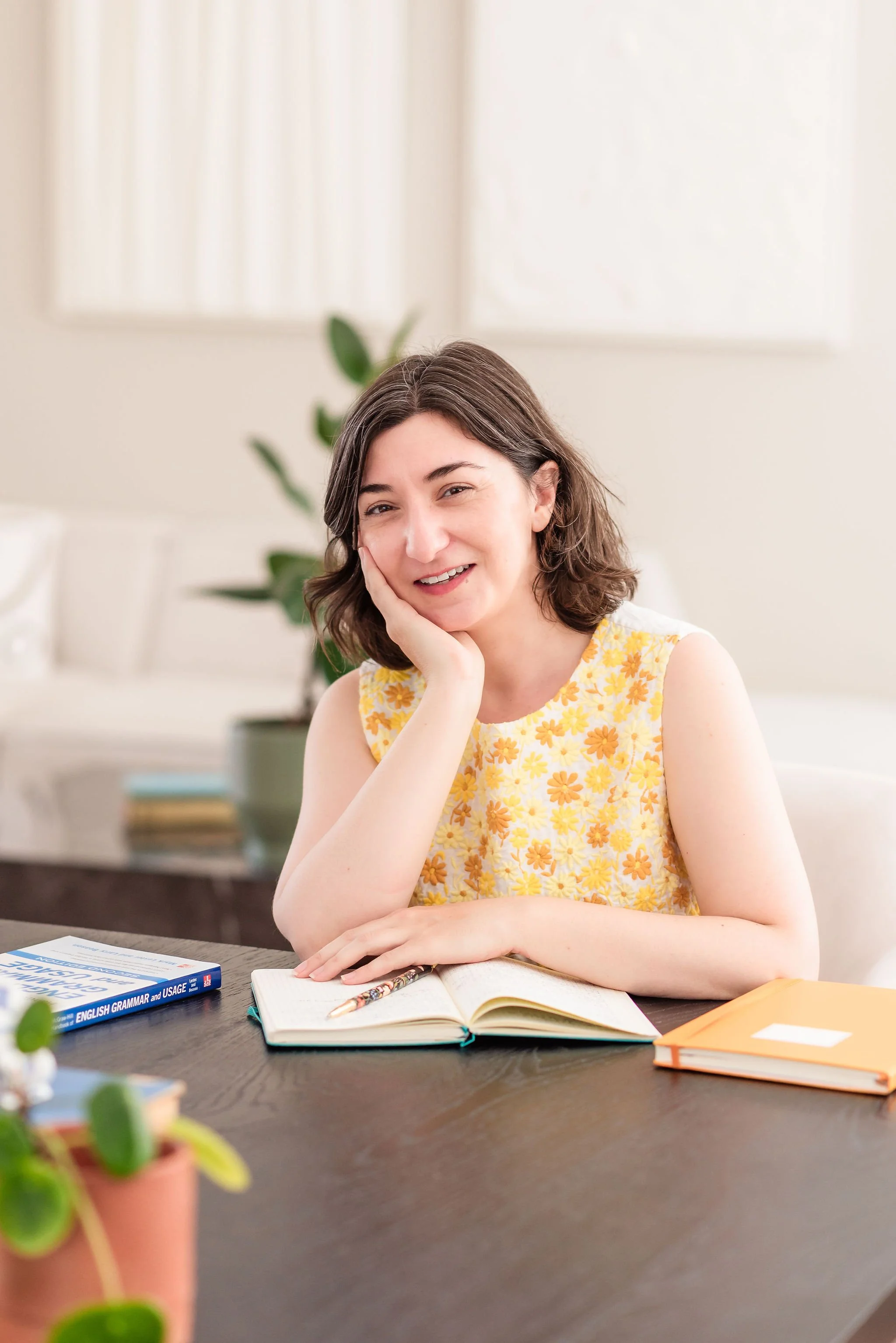 A neurodivergent woman sitting at a desk, smiling, with her face resting in one hand. There is an open notebook, a pen, and grammar book before her. She wears a yellow floral top in a bright, well-lit room with a potted plant in the background.