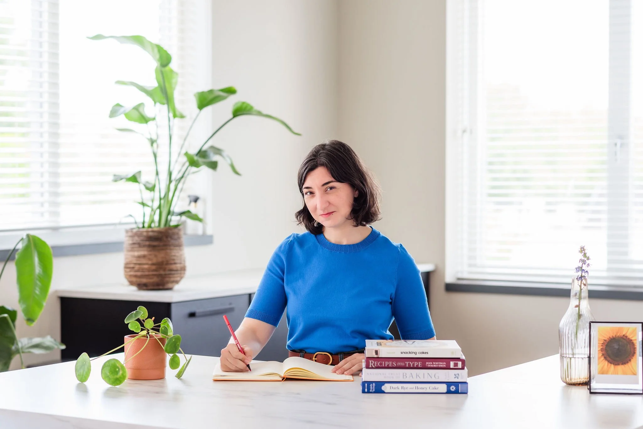 A neurodivergent woman with short brown hair and a bright blue top stands behind a white kitchen counter, smiling gently into the camera. Her pen poised on an open notebook before her, and a stack of cookbooks sits on the counter beside her.