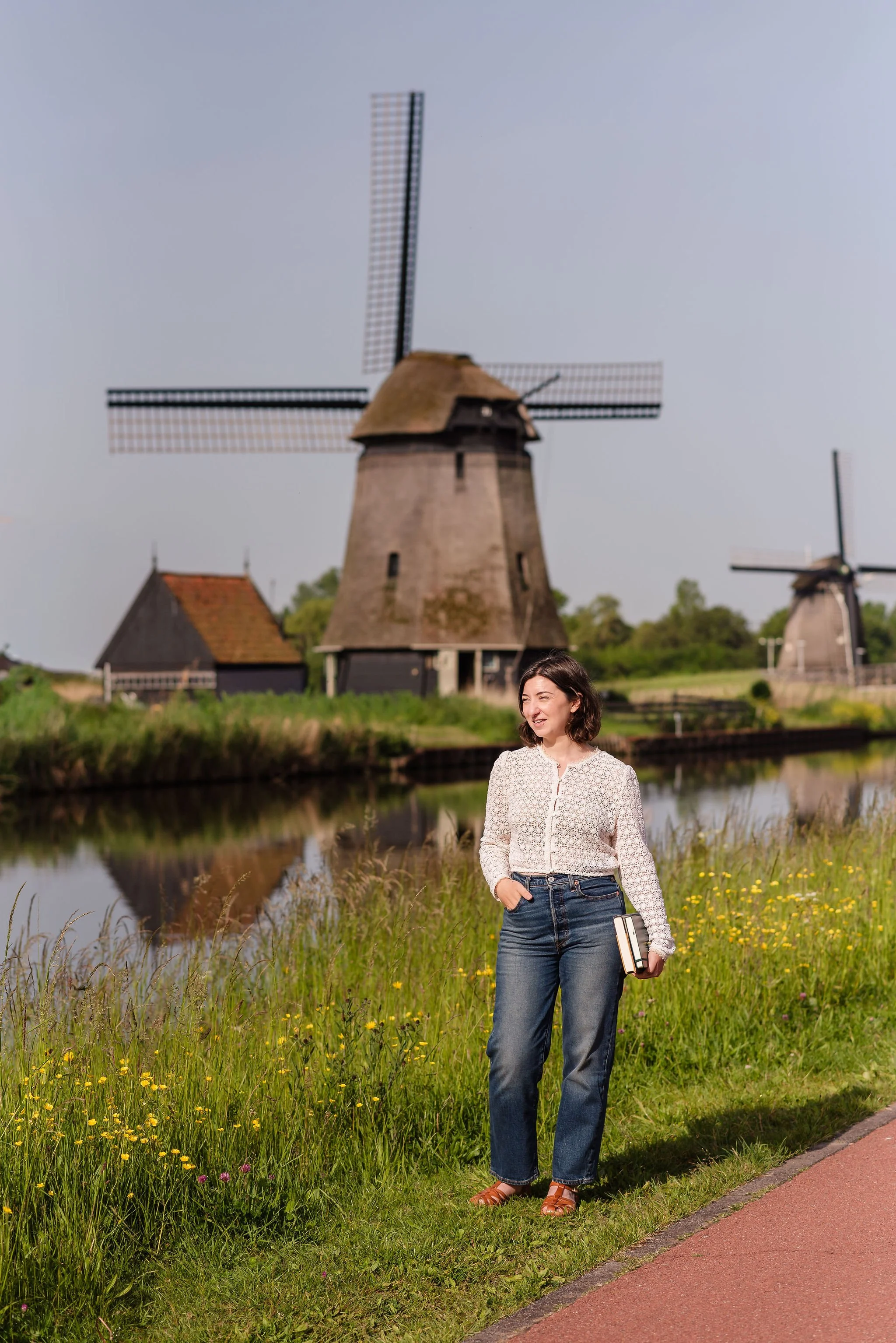 A neurodivergent woman with shoulder length brown hair standing outdoors in the sun in the Netherlands near a canal with old-fashioned windmills in the background, holding a book and looking thoughtfully into the distance.