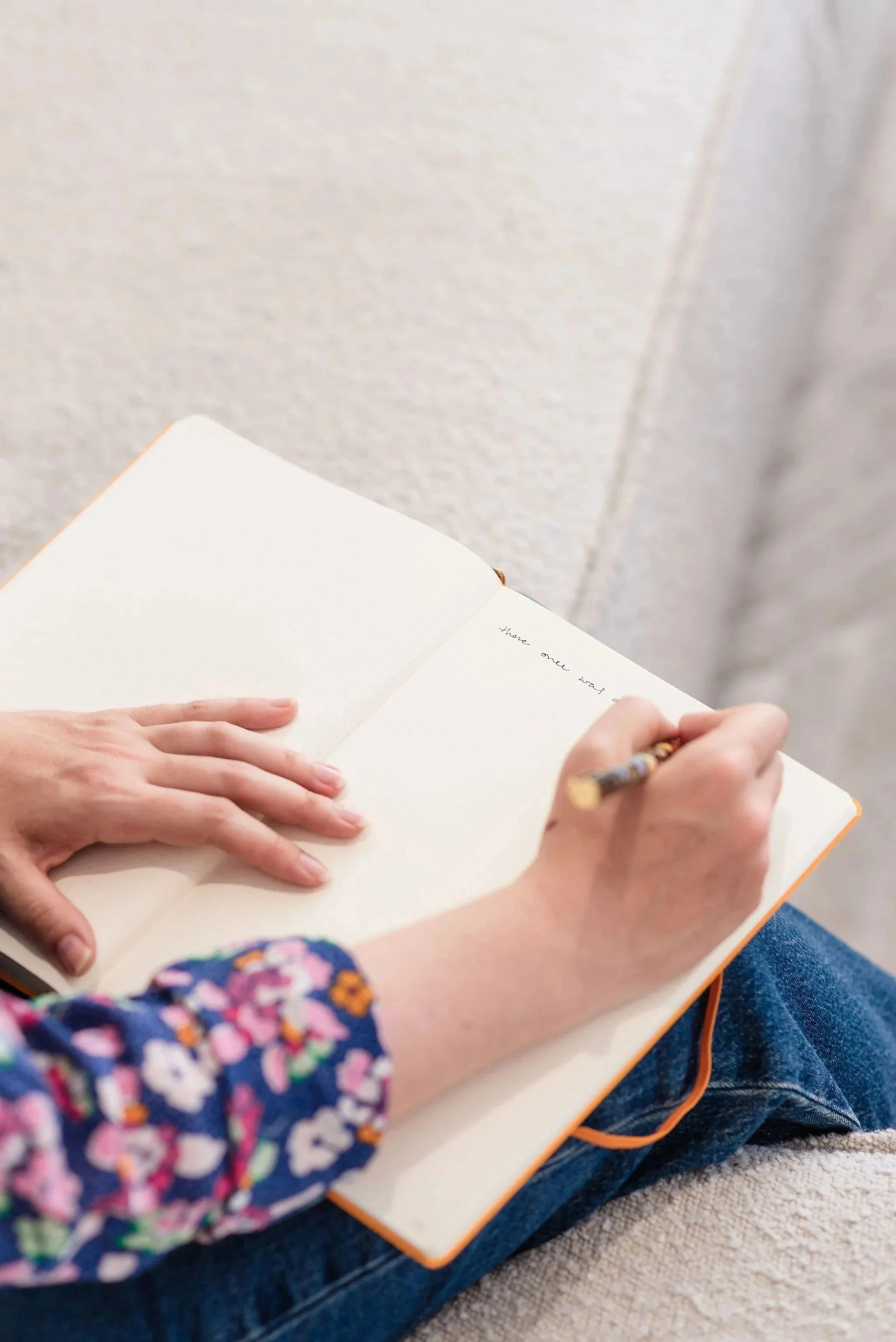Only visible are the hands forearms of a woman wearing a colorful floral blouse, as she writes in a notebook and rests her other hand on the open pages.