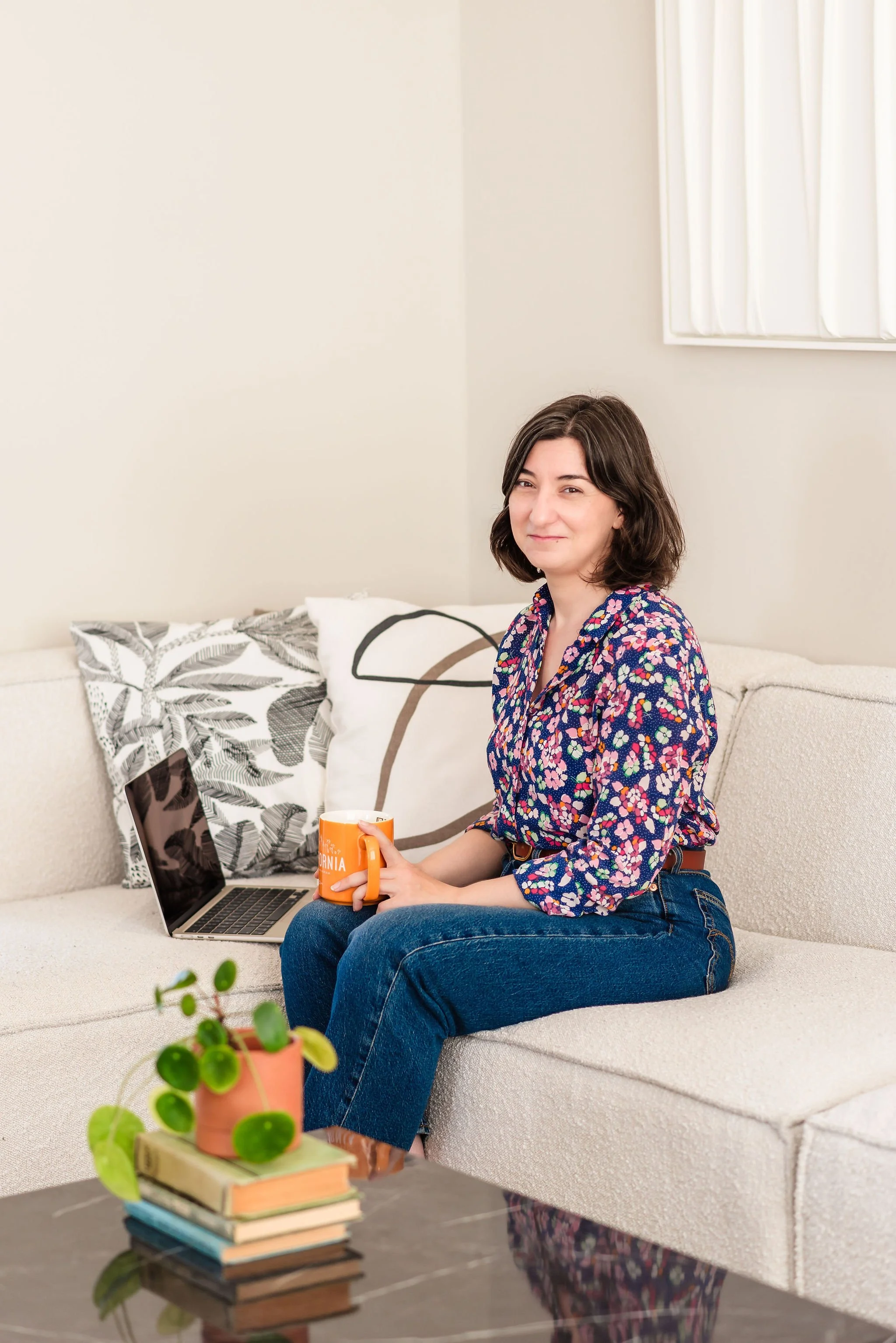 A neurodivergent woman with short brown hair and a floral shirt sitting on a beige sofa, holding an orange mug, a laptop beside her, in a room with white walls and white textural art, a stack of books and a potted plant on top in the foreground.
