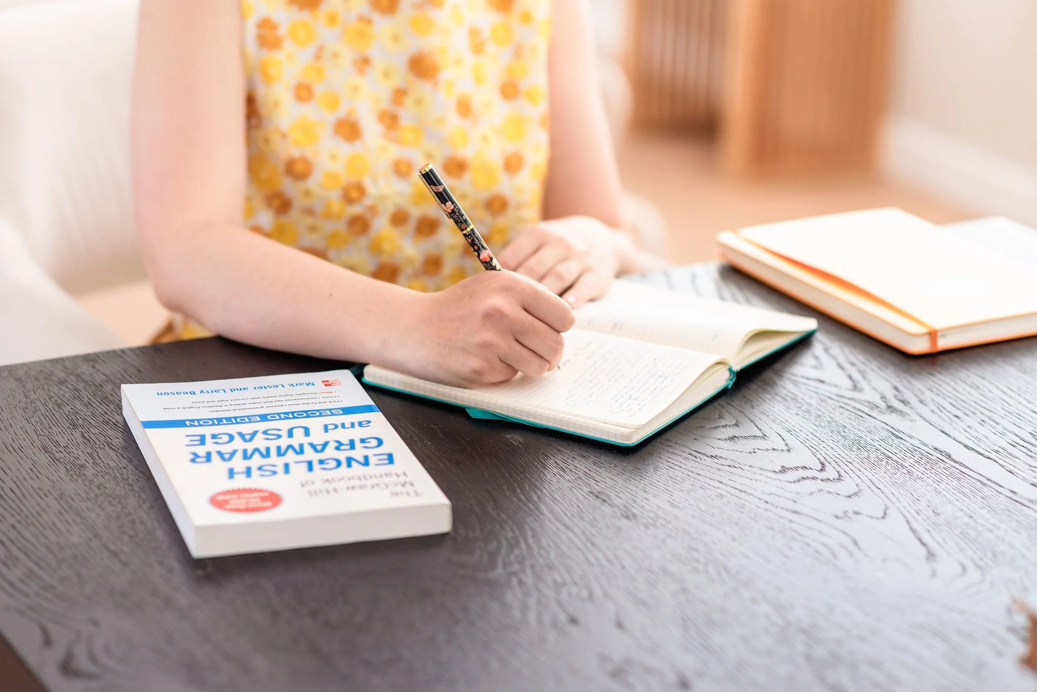 A neurodivergent woman wearing a yellow floral top sits at a table and writes in an open notebook, an English grammar book beside her. Her face is not visible.