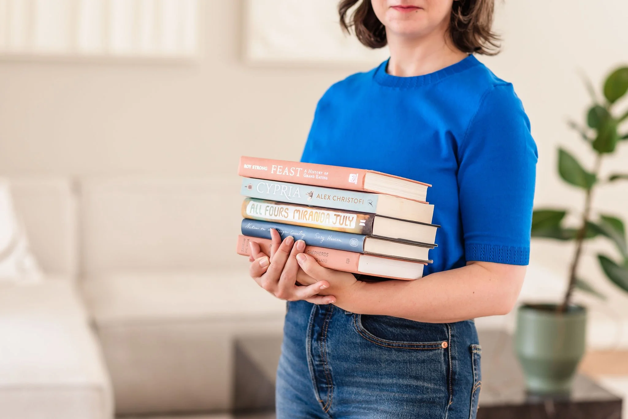 A neurodivergent woman, only her torso visible, wearing a bright blue sweater and holding a stack of five books in a room in front of her chest in a room with neutral-colored walls.