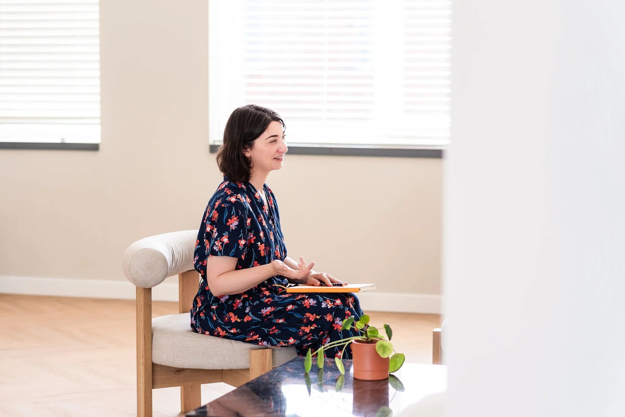 A neurodivergent woman with shoulder-length brown hair wearing a blue floral jumpsuit sits sideways to the camera on a low chair in a bright room. She is talking and smiling with a notebook in her lap. A potted plant sits in the foreground.