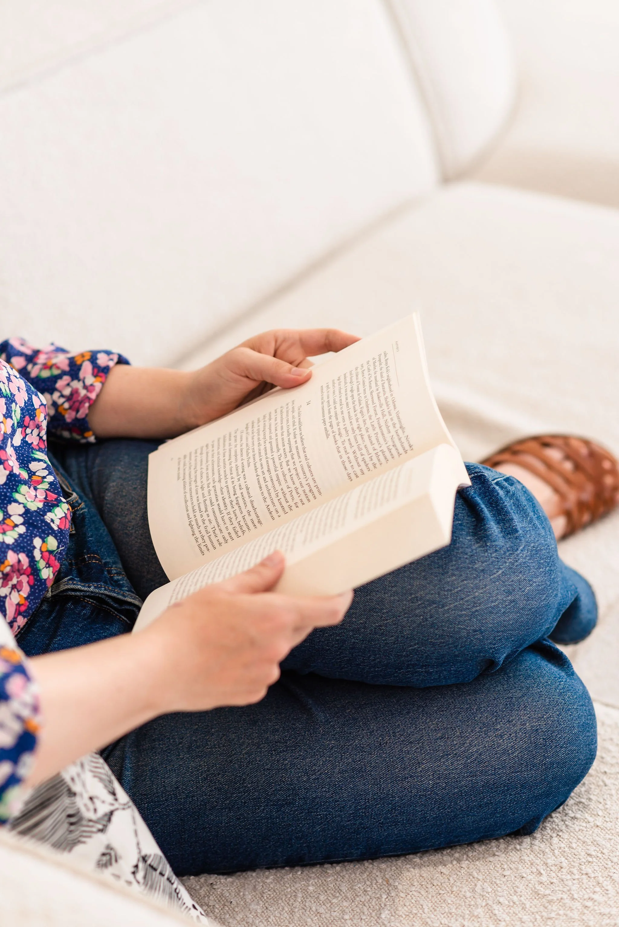 A neurodivergent woman wearing a jeans and a colorful floral blouse sits on with her knees up a couch and reads a book in her lap; her face is not visible.