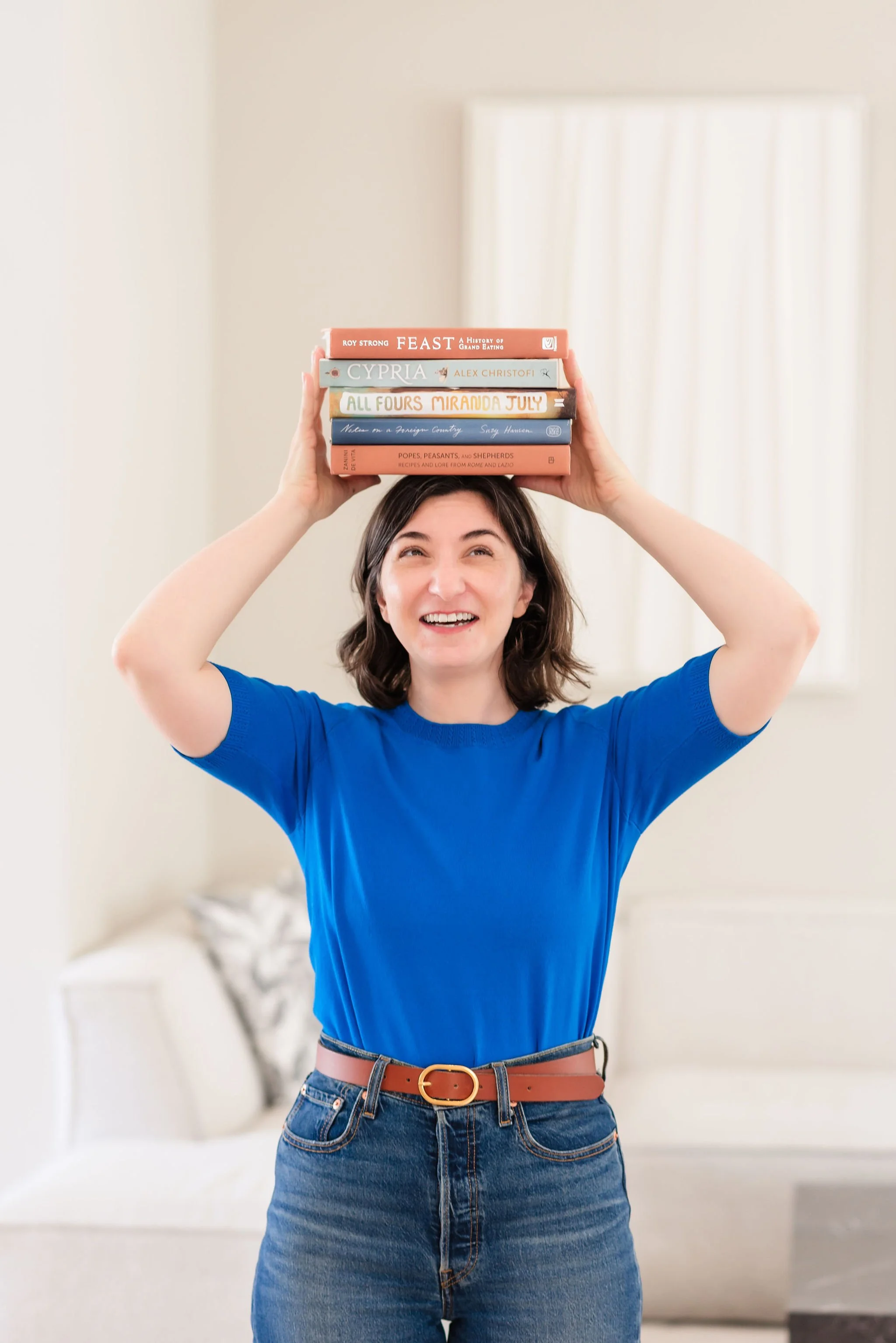 A neurodivergent woman with shoulder-length brown hair wearing a blue shirt and high-waisted jeans with a brown belt, smiling and holding a stack of five books on her head, standing in a bright, softly lit room.