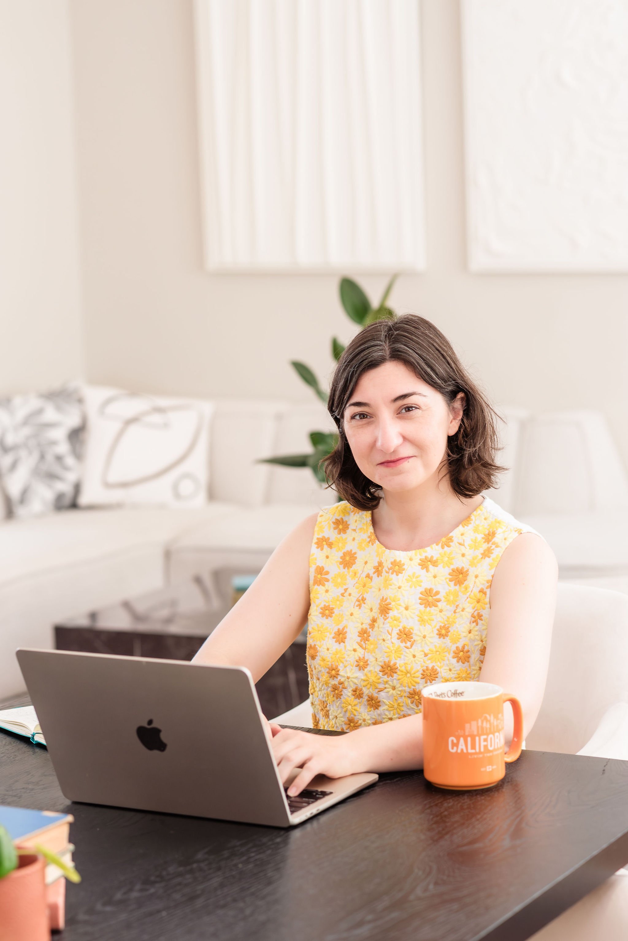 A neurodivergent woman with short brown hair wearing a yellow floral sleeveless top sitting at a desk using a silver MacBook. She looks into the camera and smiles confidently. An orange coffee mug with 'CALIFORNIA' in white text is beside her.