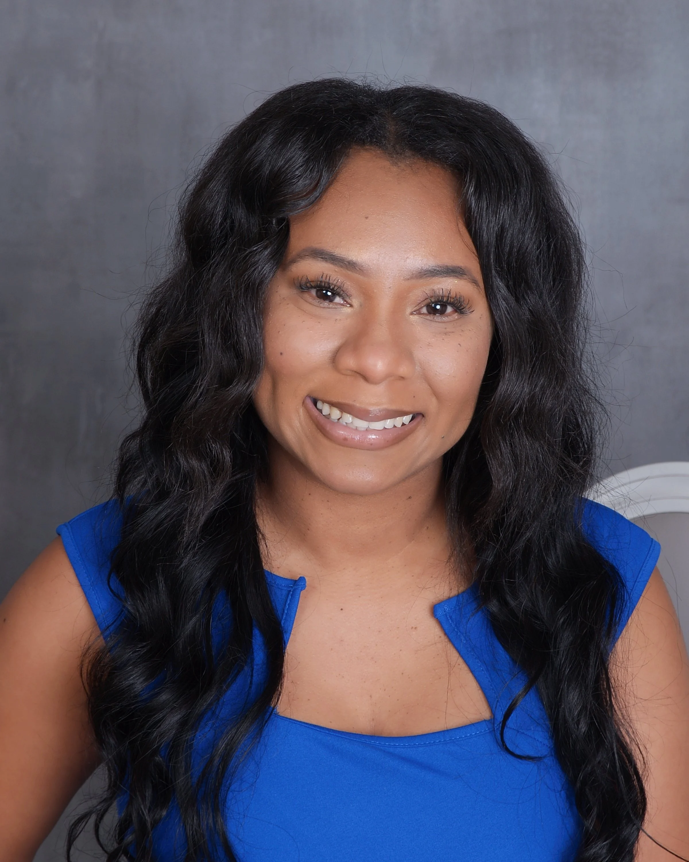 African American Therapist with long black wavy hair wearing a sleeveless blue top against a gray background.