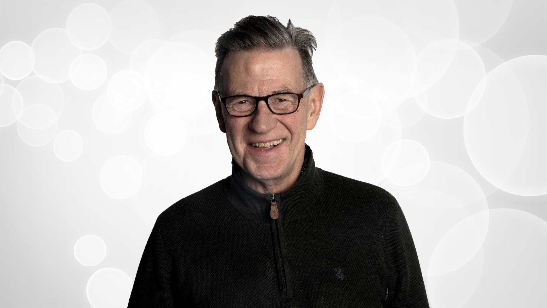 An elderly man with gray hair, glasses, smiling, wearing a black zip-up sweater, standing against a light background with circular patterns.
