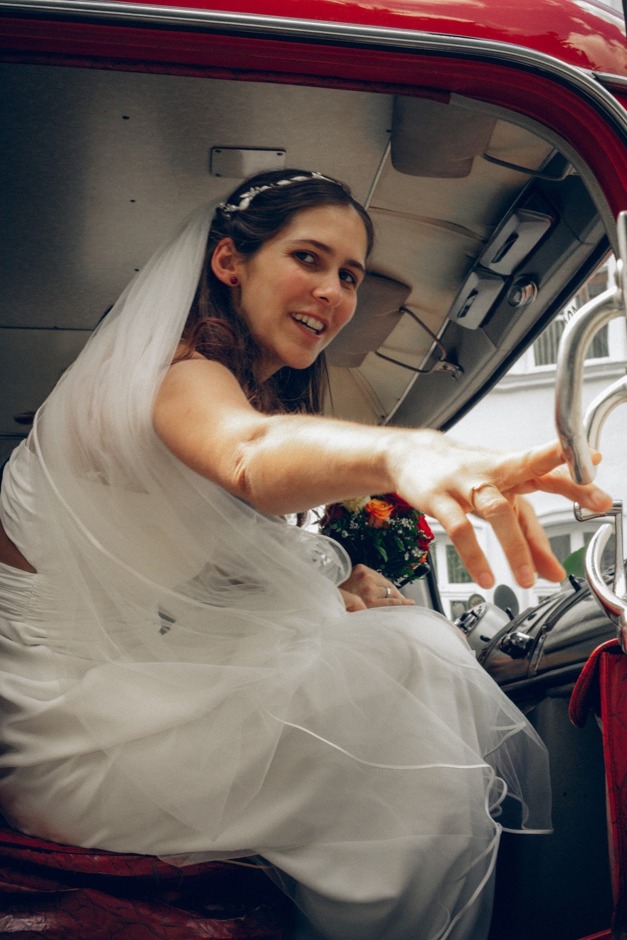 Braut in Hochzeitskleid und Schleier sitzt in einem roten Auto und lächelt.