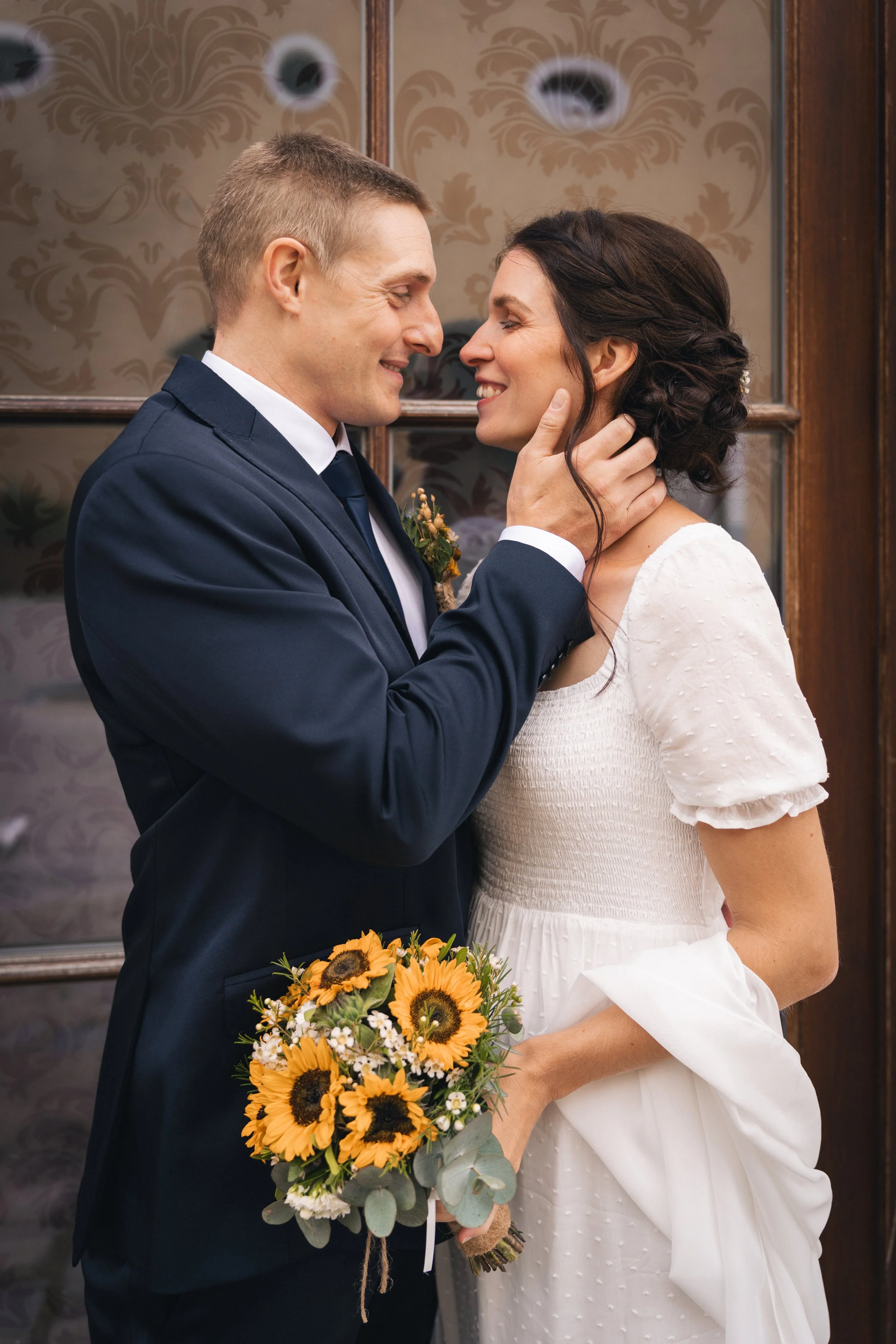 Ein Brautpaar bei einer Hochzeit, der Bräutigam hält die Braut im Arm und berührt ihr Gesicht, beide lächeln sich an, die Braut hält einen Sonnenblumenstrauß.