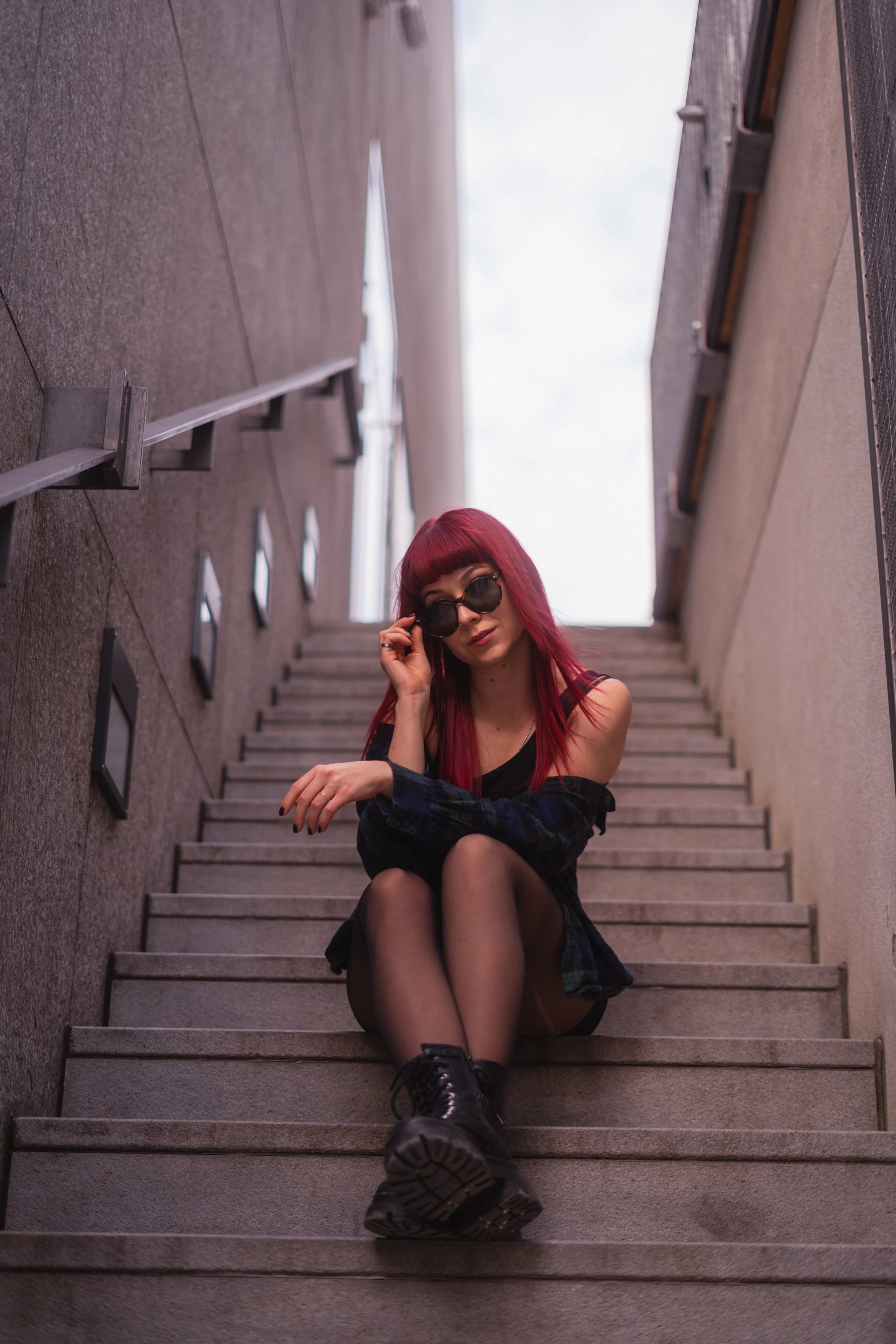 Junge Frau mit roten Haaren, Sonnenbrille, schwarzem Top, off-shoulder Hemd, schwarzen Strumpfhosen und schwarzen Stiefeln sitzt auf Treppe zwischen modernen Gebäuden im Freien.