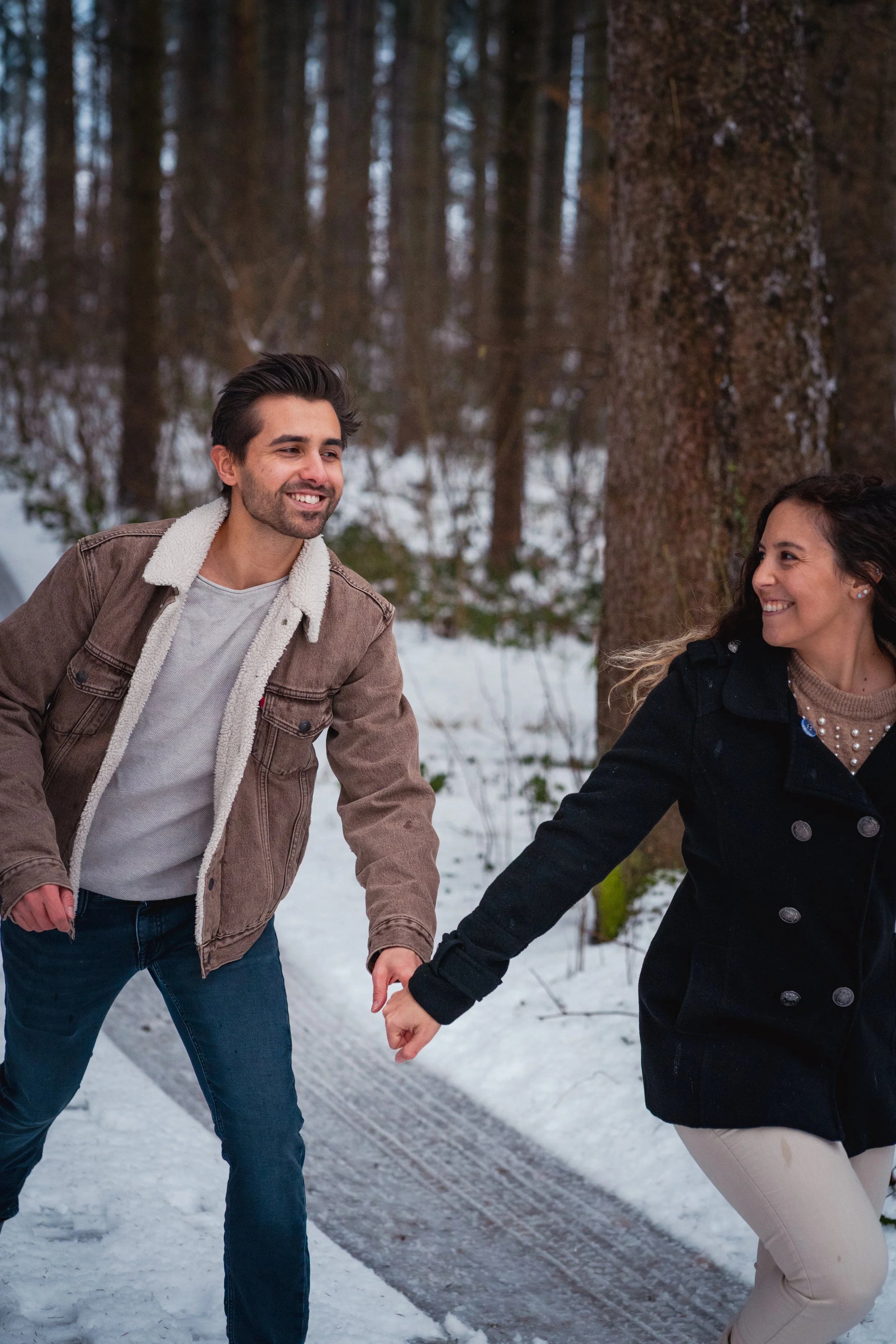 Ein lachendes Paar, das im Schnee im Wald spazieren geht und sich hält.