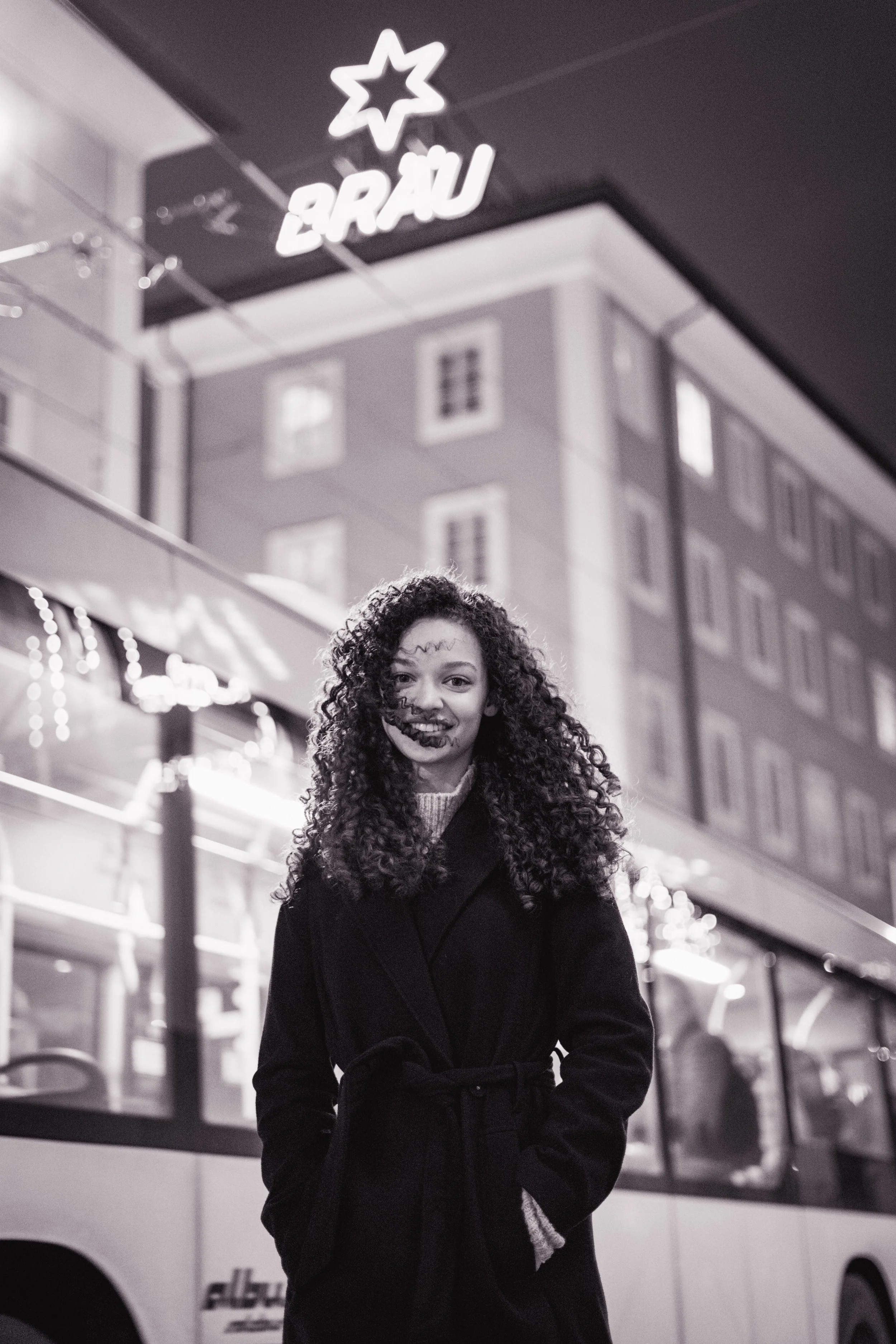 Eine junge Frau mit lockigem Haar trägt einen dunklen Mantel und steht vor einem Bus in einer Stadt bei Nacht. Über ihr leuchtet ein Neon-Schild mit einem Stern und der Schrift "BRÄU".