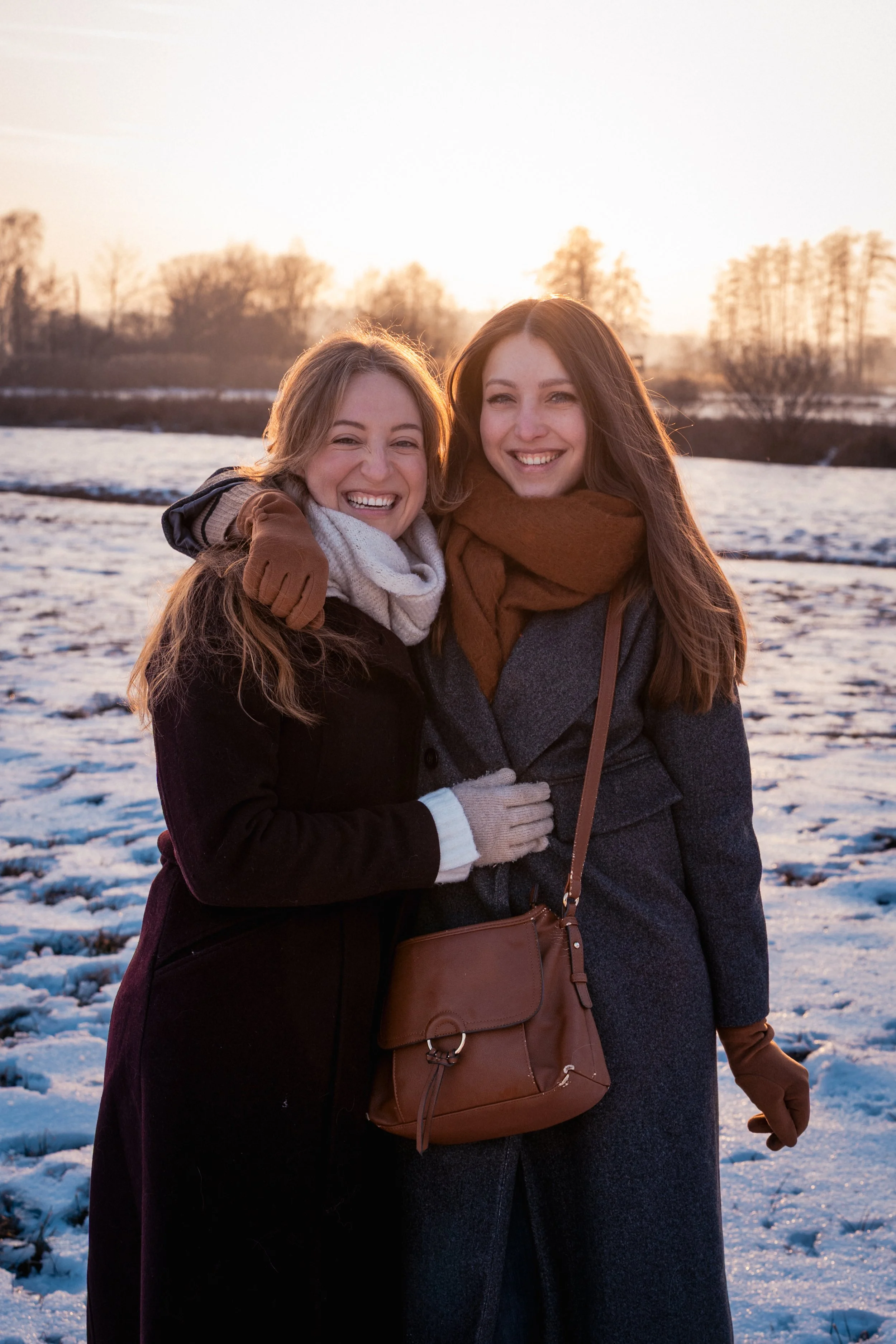 Zwei lachende Frauen im Winter draußen, umgeben von verschneitem Feld und Bäumen bei Sonnenuntergang.
