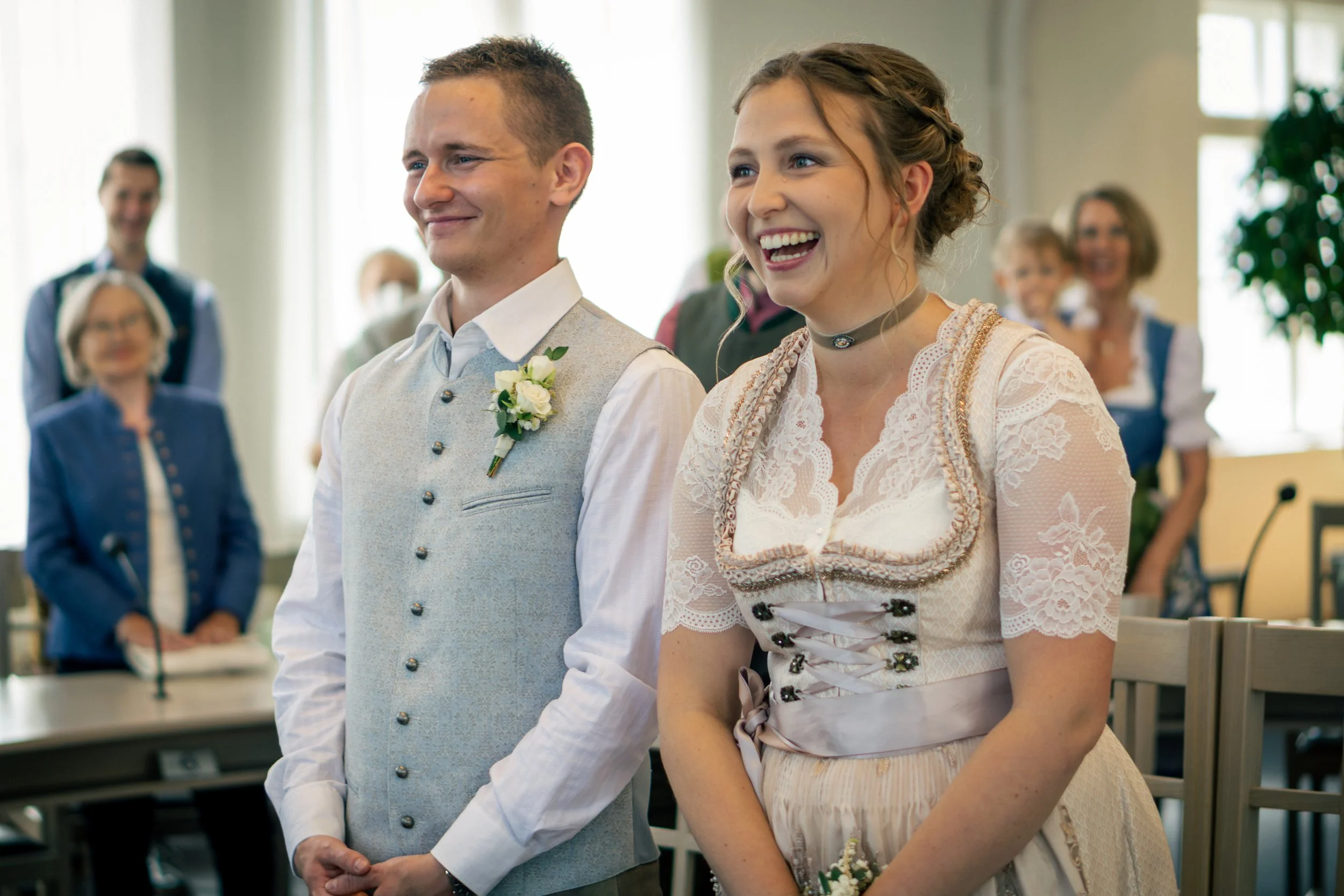 Brautpaar bei einer Hochzeit, lachend und umgeben von Gästen in traditionellen Trachten.