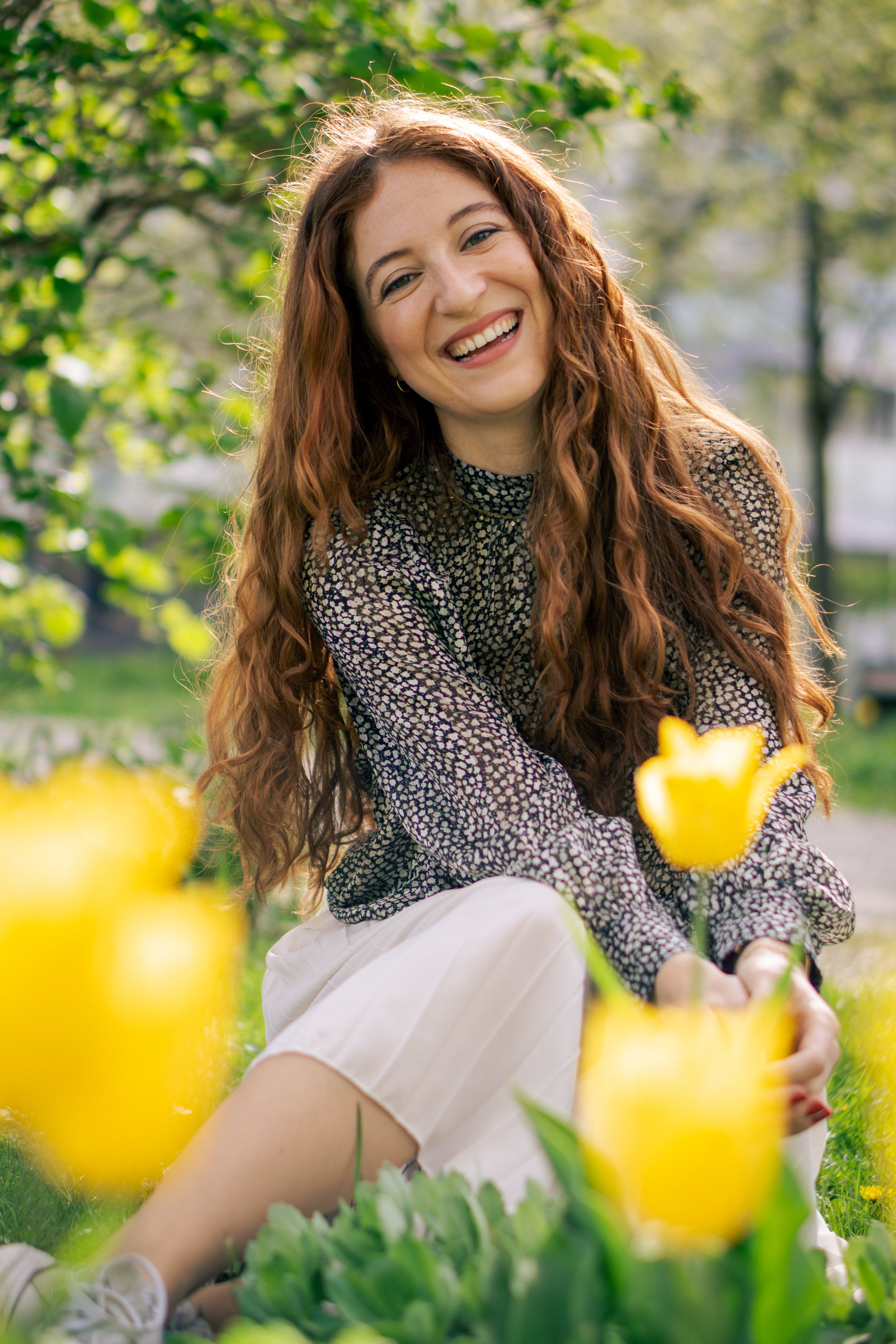 Lächelnde Frau mit lockigem braunem Haar sitzt im Garten unter Bäumen, umgeben von gelben Tulpen