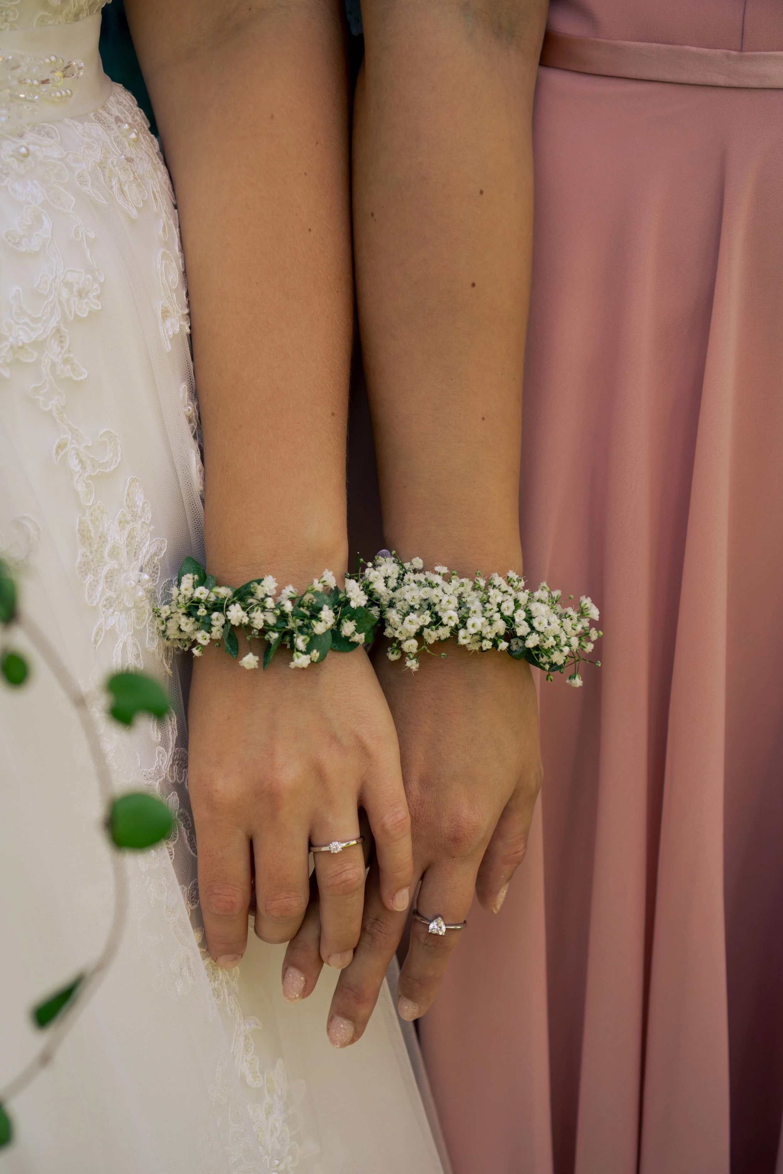 Hände mit Eheringen, Blumenarmbändern und Brautkleid sowie Brautjungfernkleid im Hintergrund.