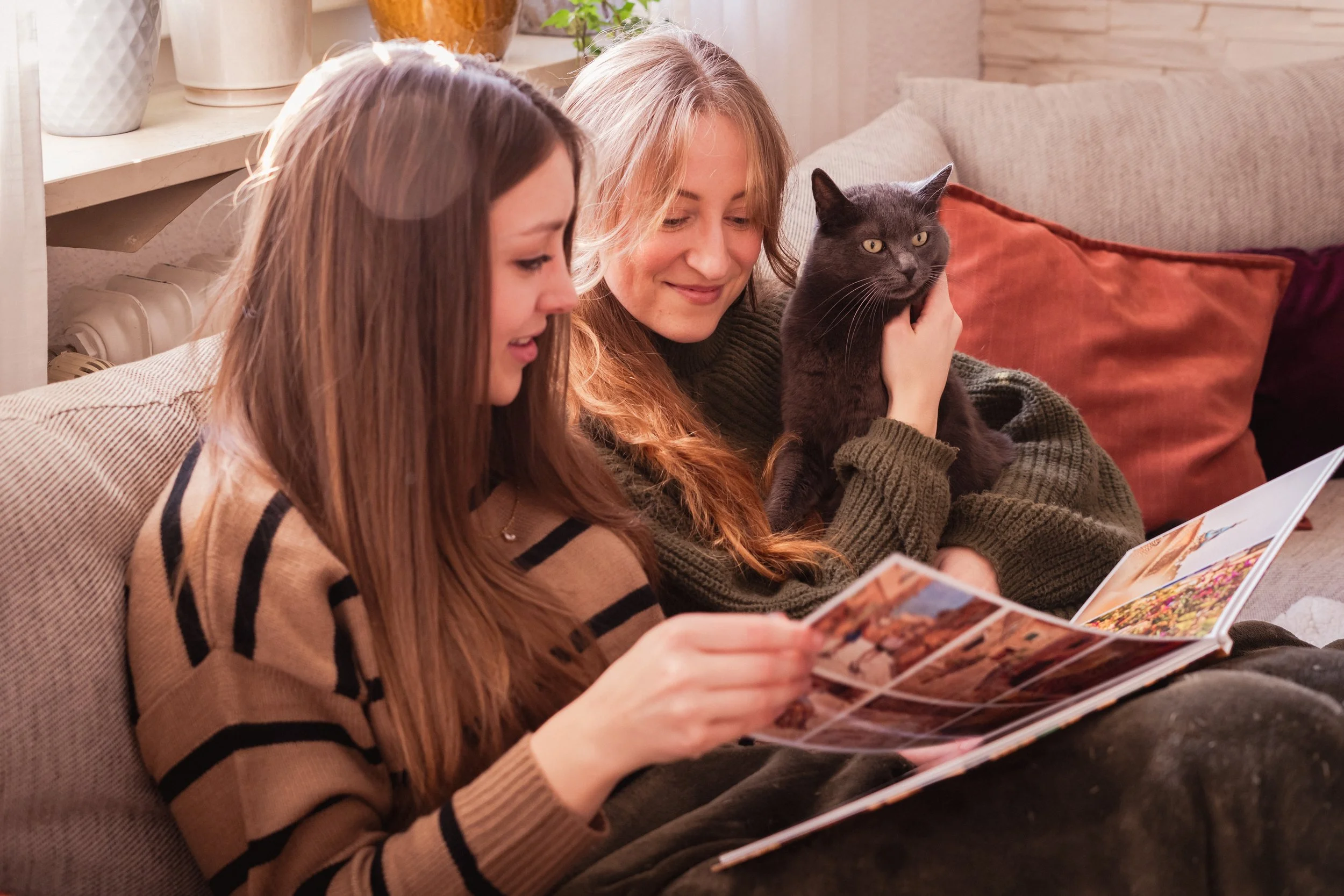 Zwei Frauen sitzen auf einem Sofa, eine liest eine Zeitschrift, die andere hält einen schwarzen Kater in den Armen, im Hintergrund Kissen und Pflanzen.