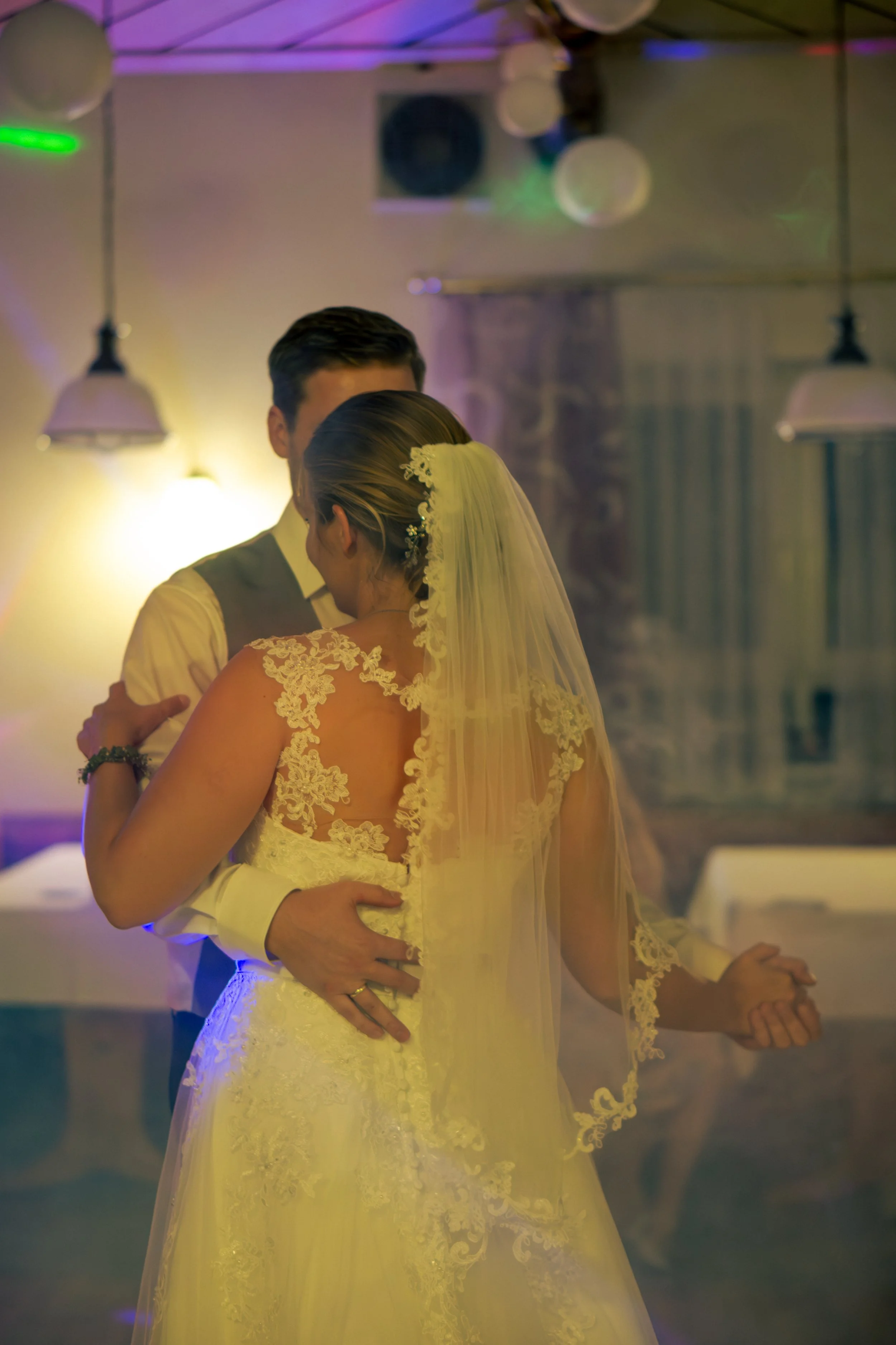 Ein Brautpaar tanzt auf einer Hochzeit im festlich dekorierten Raum, die Braut trägt ein elegantes weißes Kleid mit Spitze und Schleier, der Bräutigam trägt einen Anzug. Die Beleuchtung ist warm mit bunten Lichteffekten.