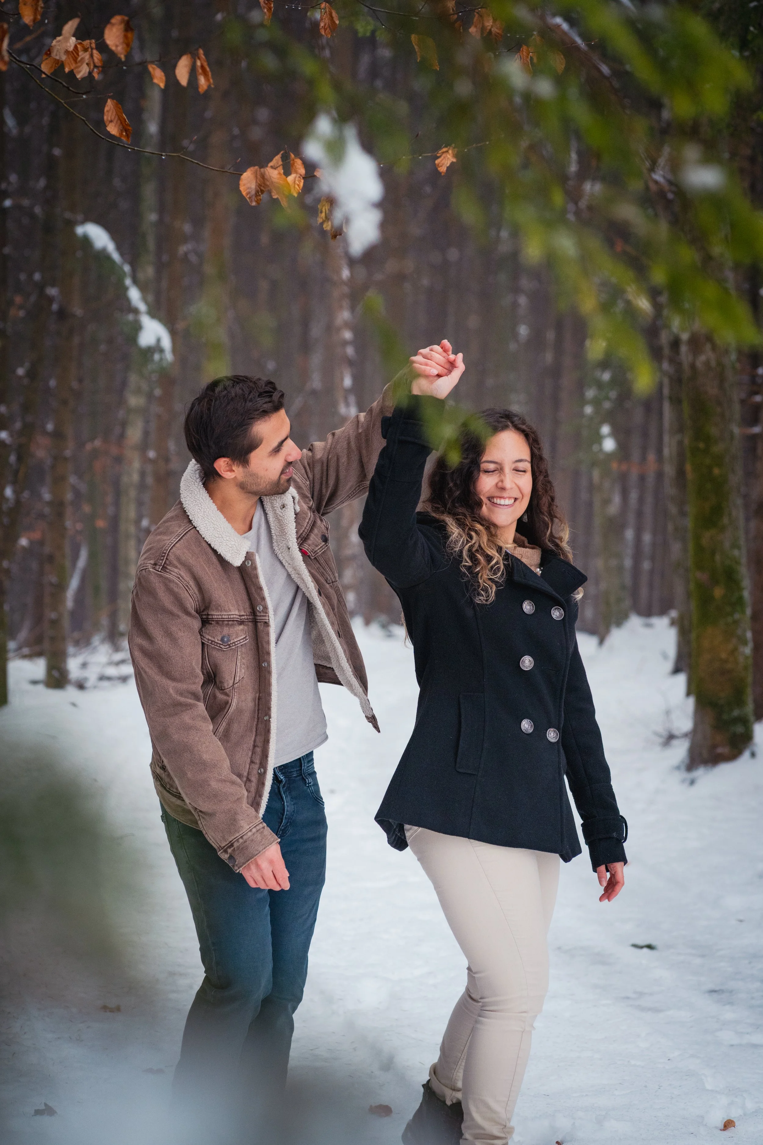 Ein Mann und eine Frau tanzen im Winterwald, während Schnee auf dem Boden liegt. Der Mann hält die Hand der Frau, und beide lachen glücklich.