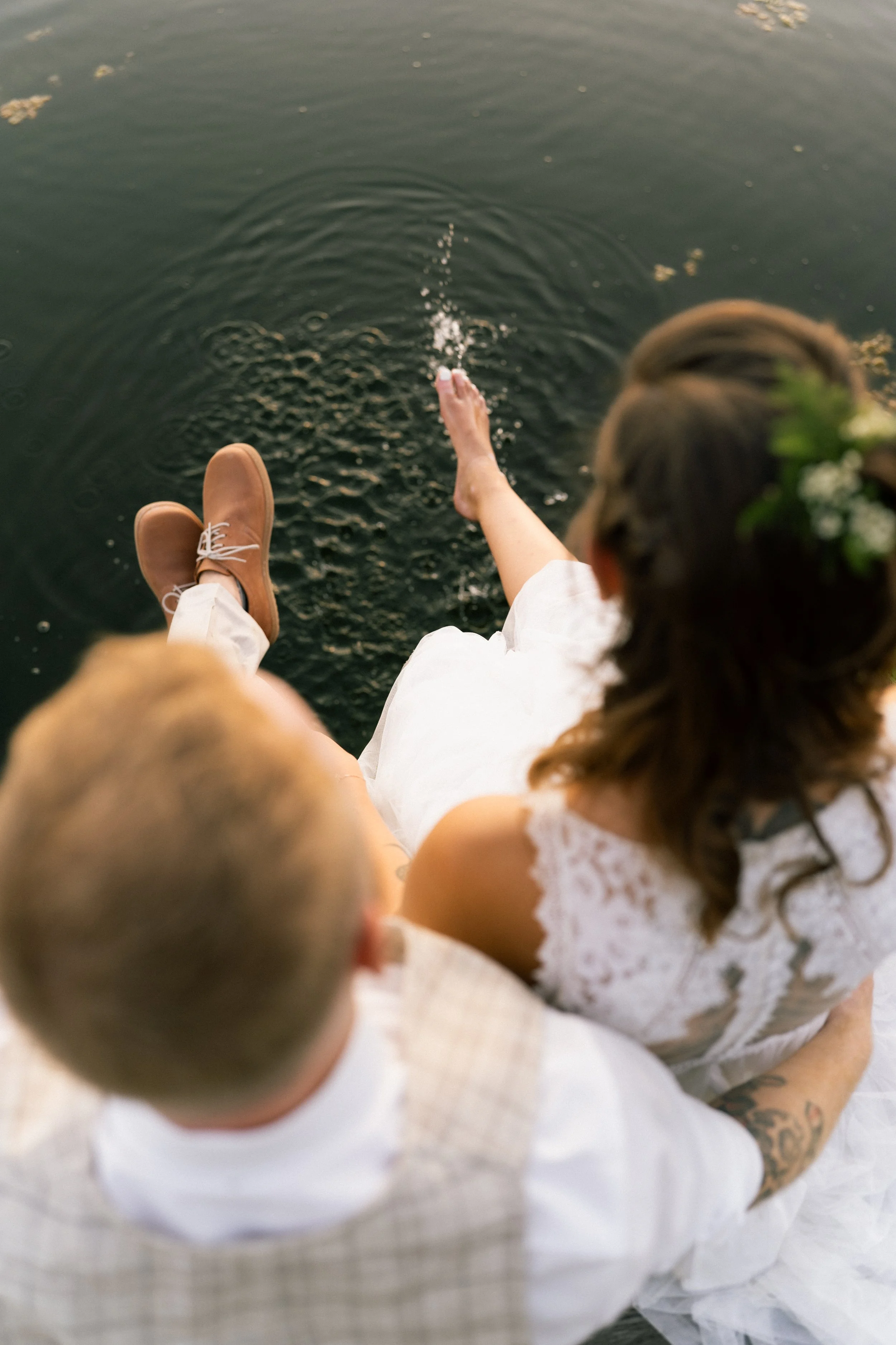 Eine Frau in einem weißen Kleid sitzt am Wasser und streckt ihre Hand ins Wasser, während ein Mann neben ihr sitzt in Linz. Die Szene wirkt entspannt und natürlich.
