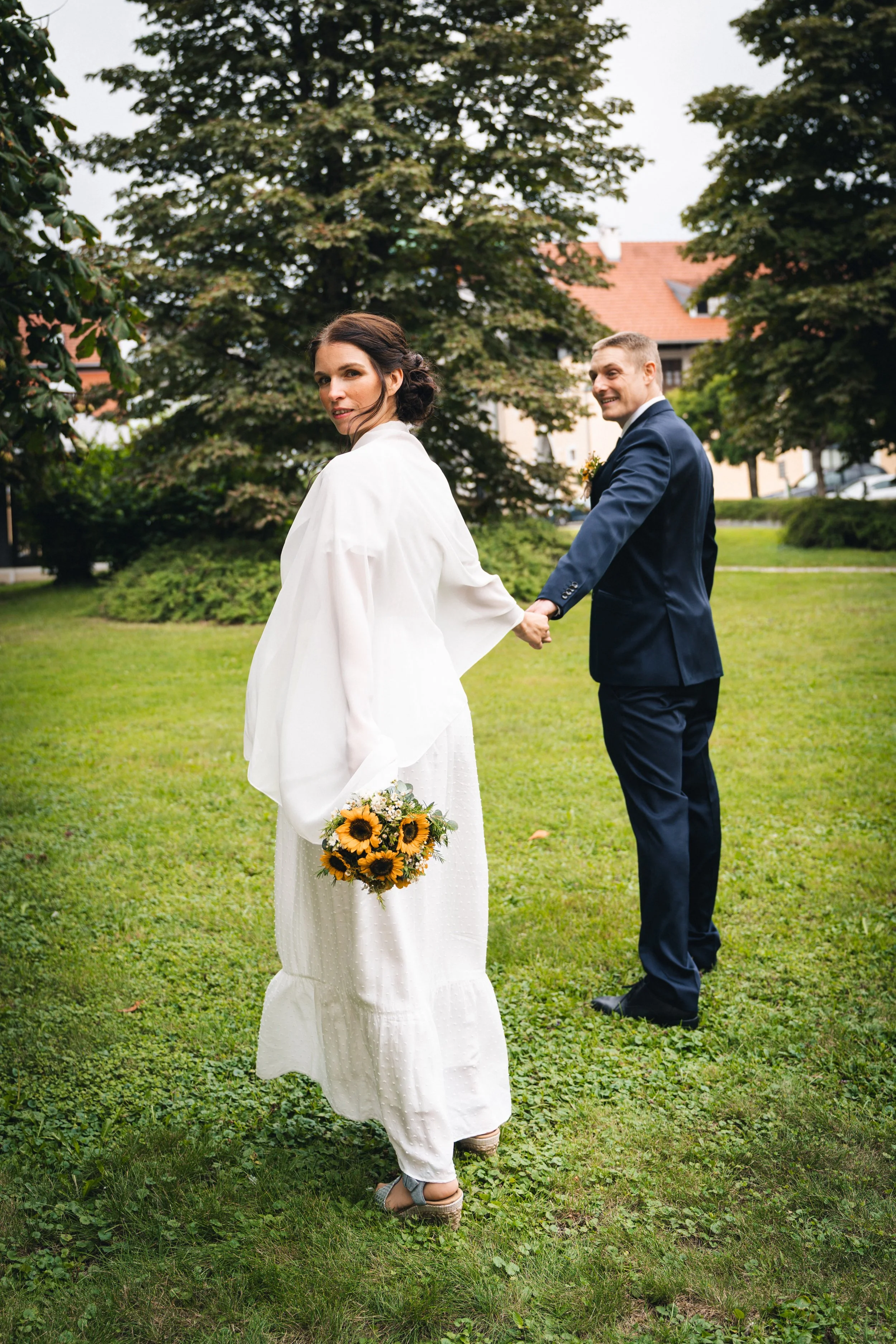 Ein Brautpaar geht Hand in Hand durch einen Garten, die Braut trägt ein langes weißes Kleid und hält einen Sonnenblumenstrauß, während der Bräutigam in einem dunklen Anzug lächelnd zuschaut.