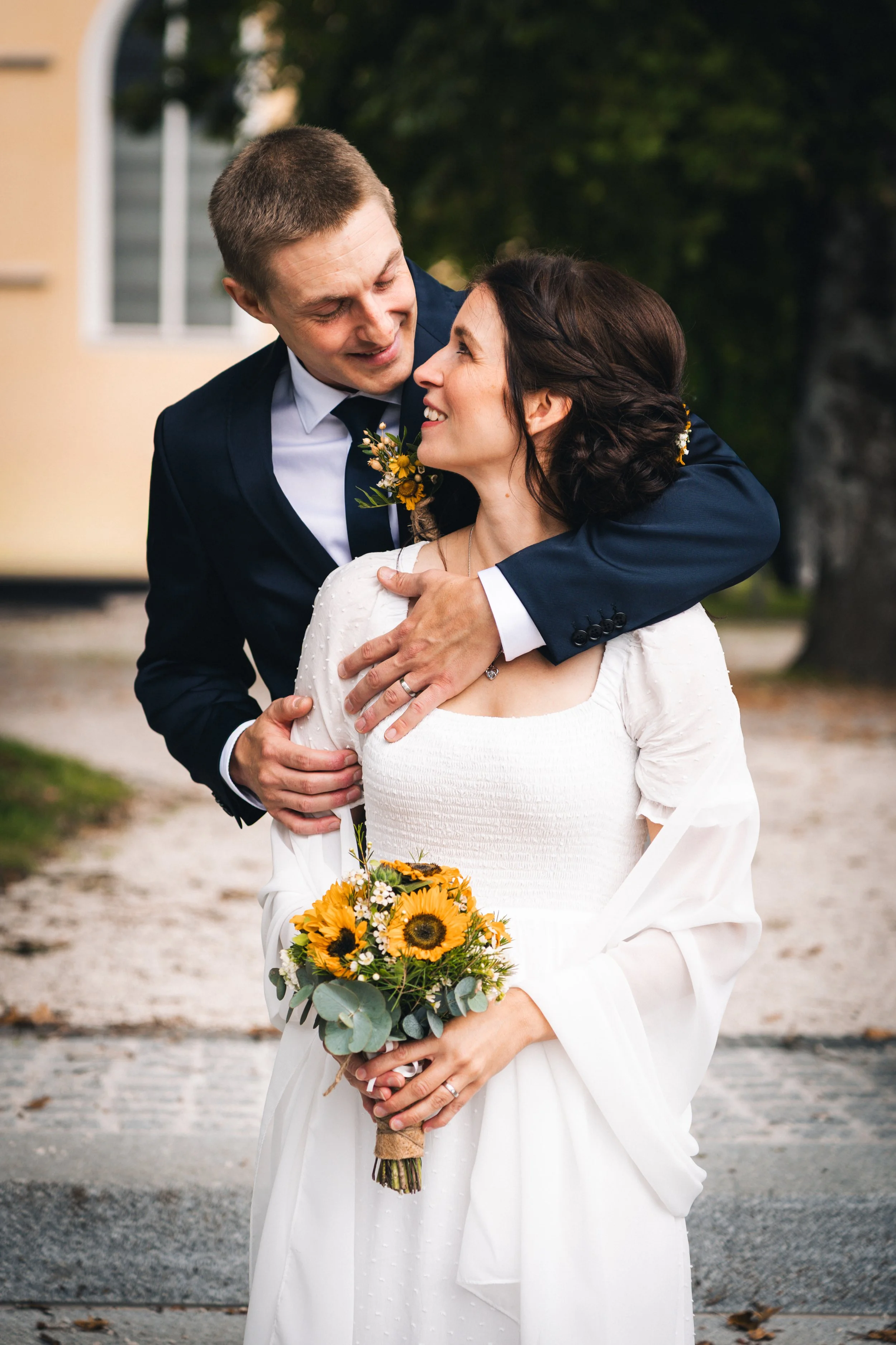 Ein Brautpaar bei einer Hochzeit, der Bräutigam umarmt die Braut und beide lächeln sich glücklich an, die Braut hält einen Sonnenblumenstrauß.