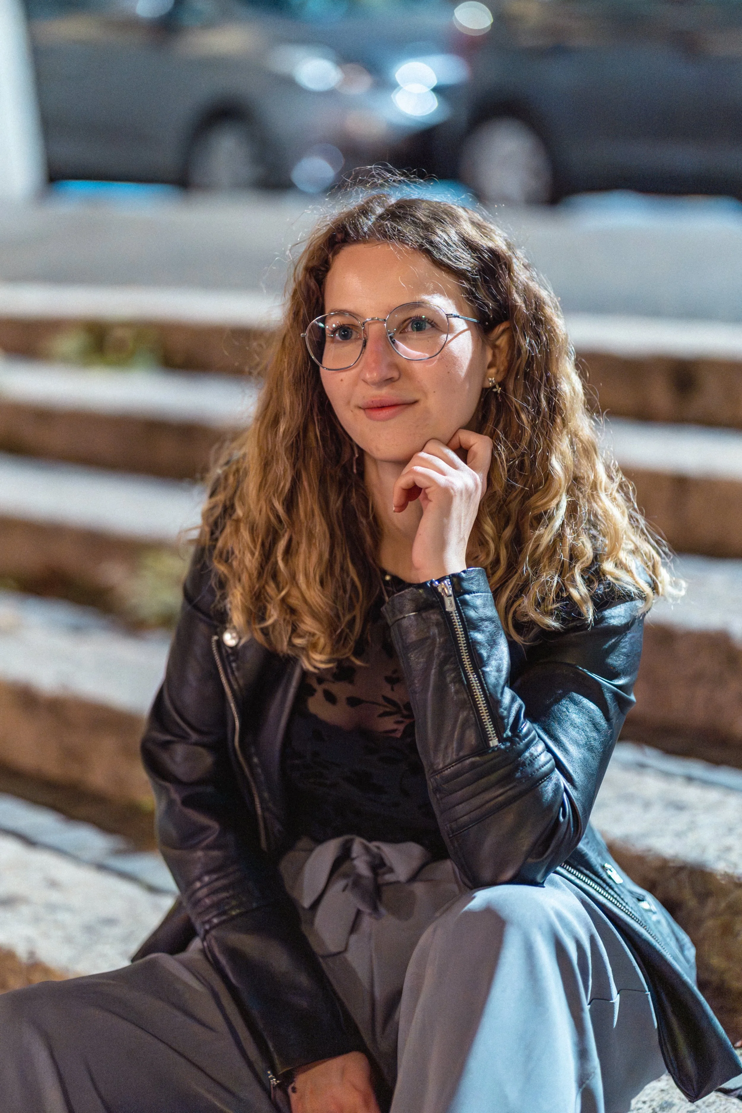 Junge Frau mit Brille und lockigem Haar, sitzend auf eine Treppe, trägt eine schwarze Lederjacke und graue Hose, im abendlichen Lichte, im Hintergrund geparkte Autos.