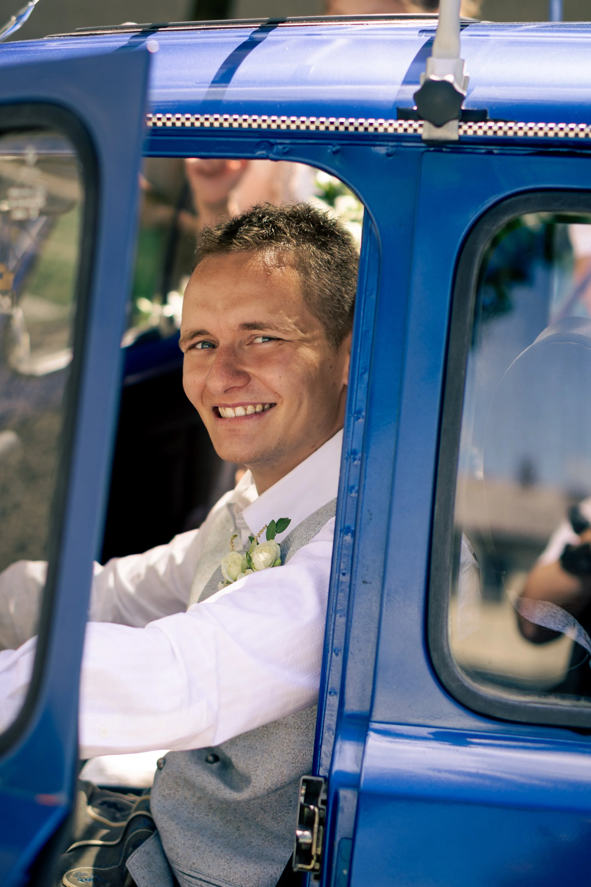 Junger Mann im weißen Hemd und Weste sitzt in einem blauen Auto, lächelt und trägt eine kleine weiße Blume am Revers.
