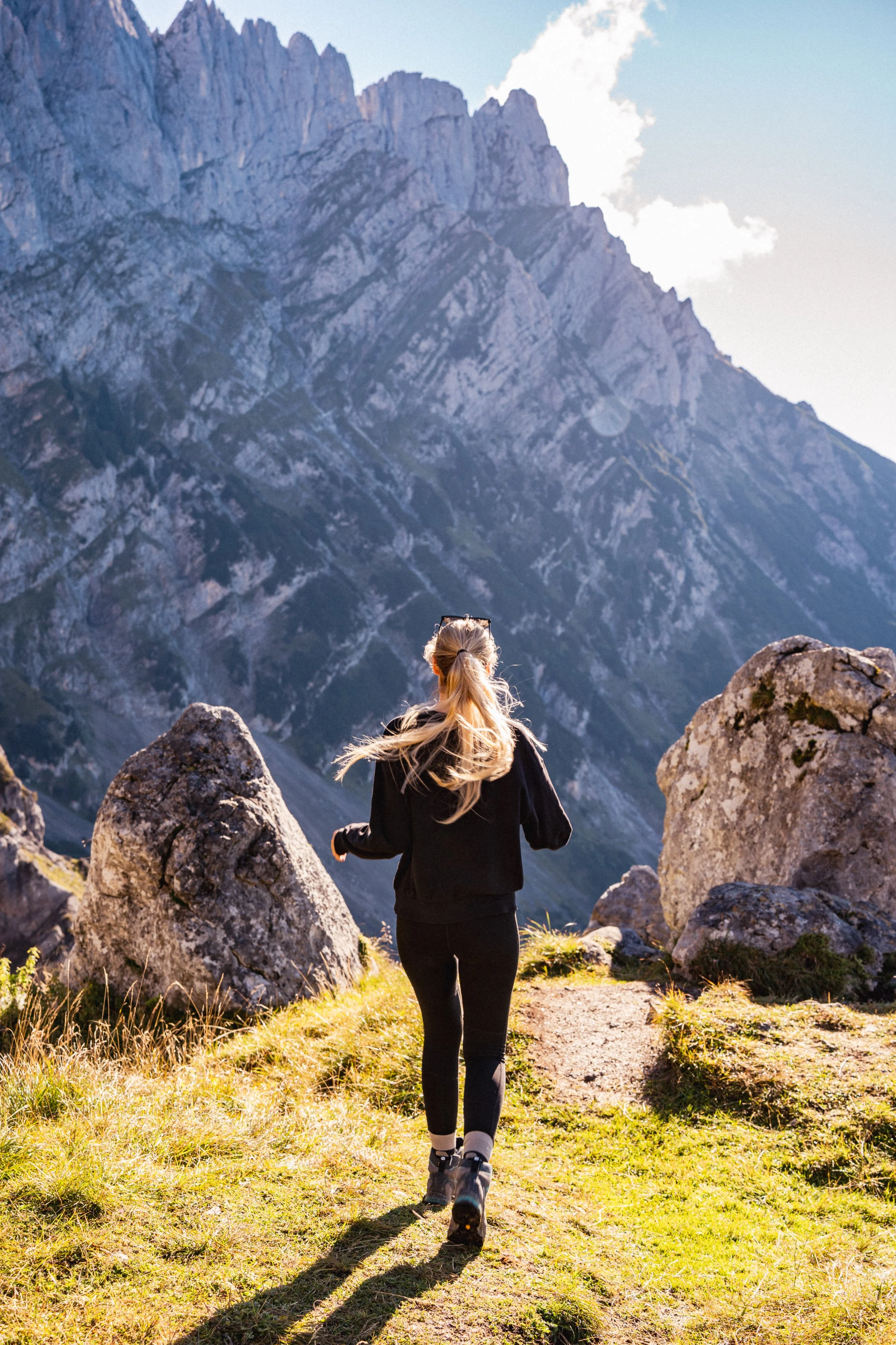Eine Frau, die in den Bergen wandert, umgeben von großen Felsen und steilen, schroffen Bergen im Hintergrund an einem sonnigen Tag.