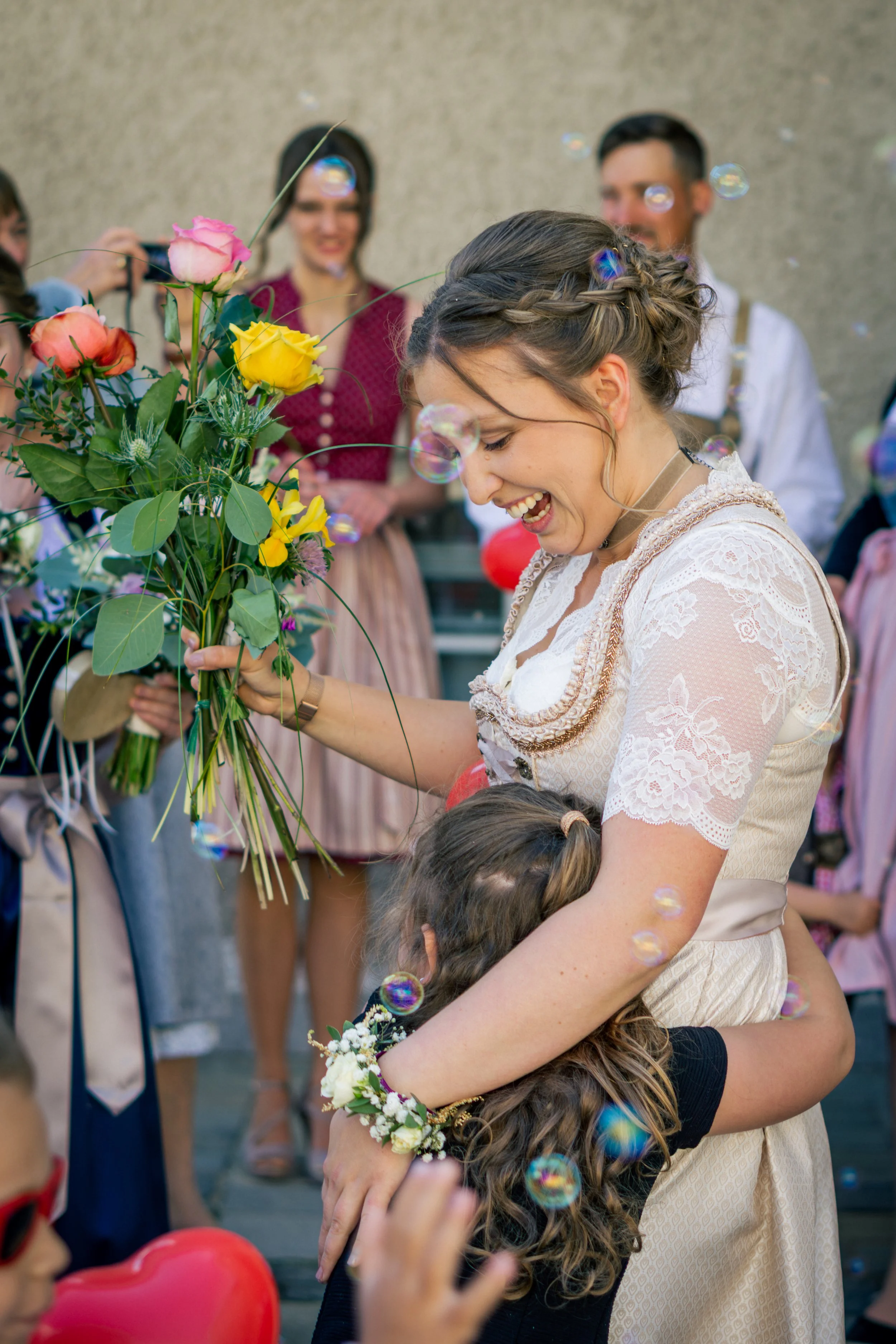 Eine lachende Frau in einem traditionellen Dirndl umarmt ein kleines Mädchen bei einer Feier, umgeben von Freunden und Blasen im Hintergrund.