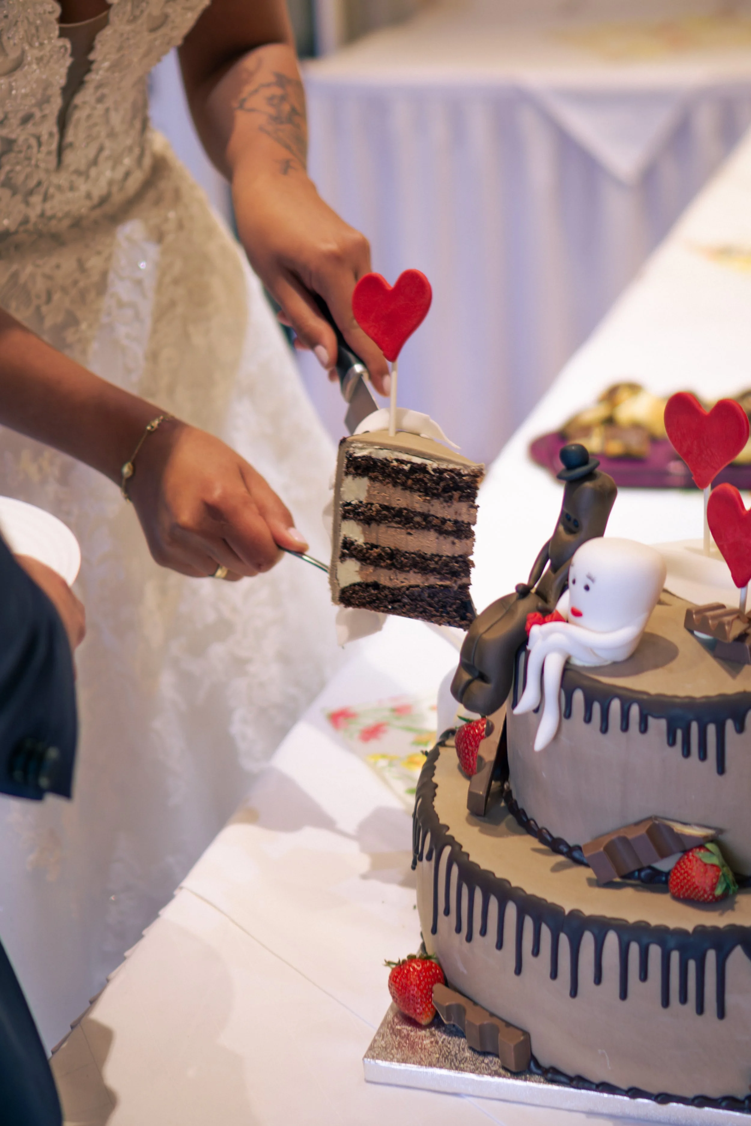 Person schneidet eine Hochzeitstorte mit mehreren Ebenen. Die Torte ist mit Schokolade und Erdbeeren dekoriert und hat Herz- und Figur-Topper.