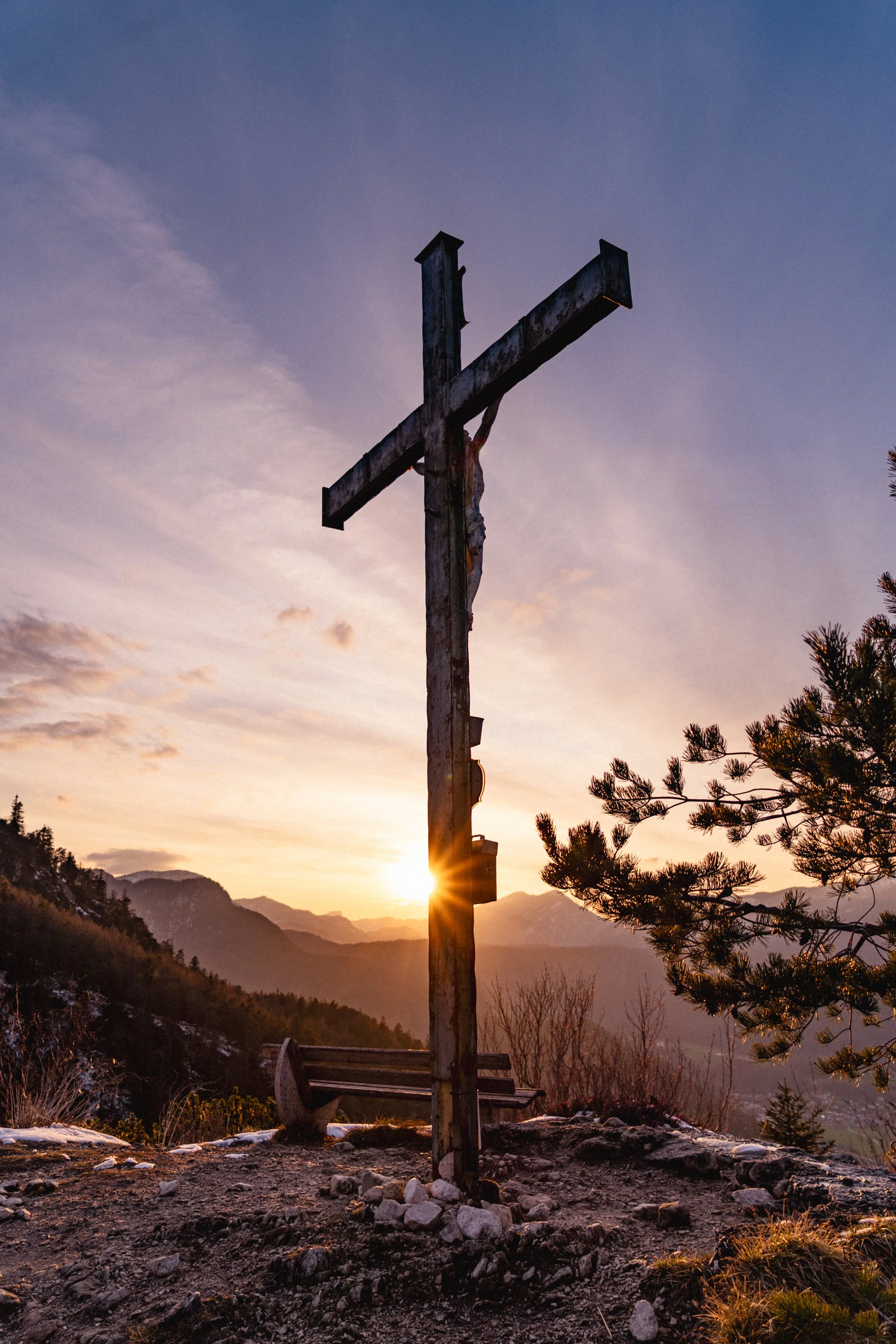 Ein Holz-Kreuz auf einem Berggipfel bei Sonnenuntergang, mit Bergketten im Hintergrund und einem schneebedeckten Baum auf der rechten Seite.