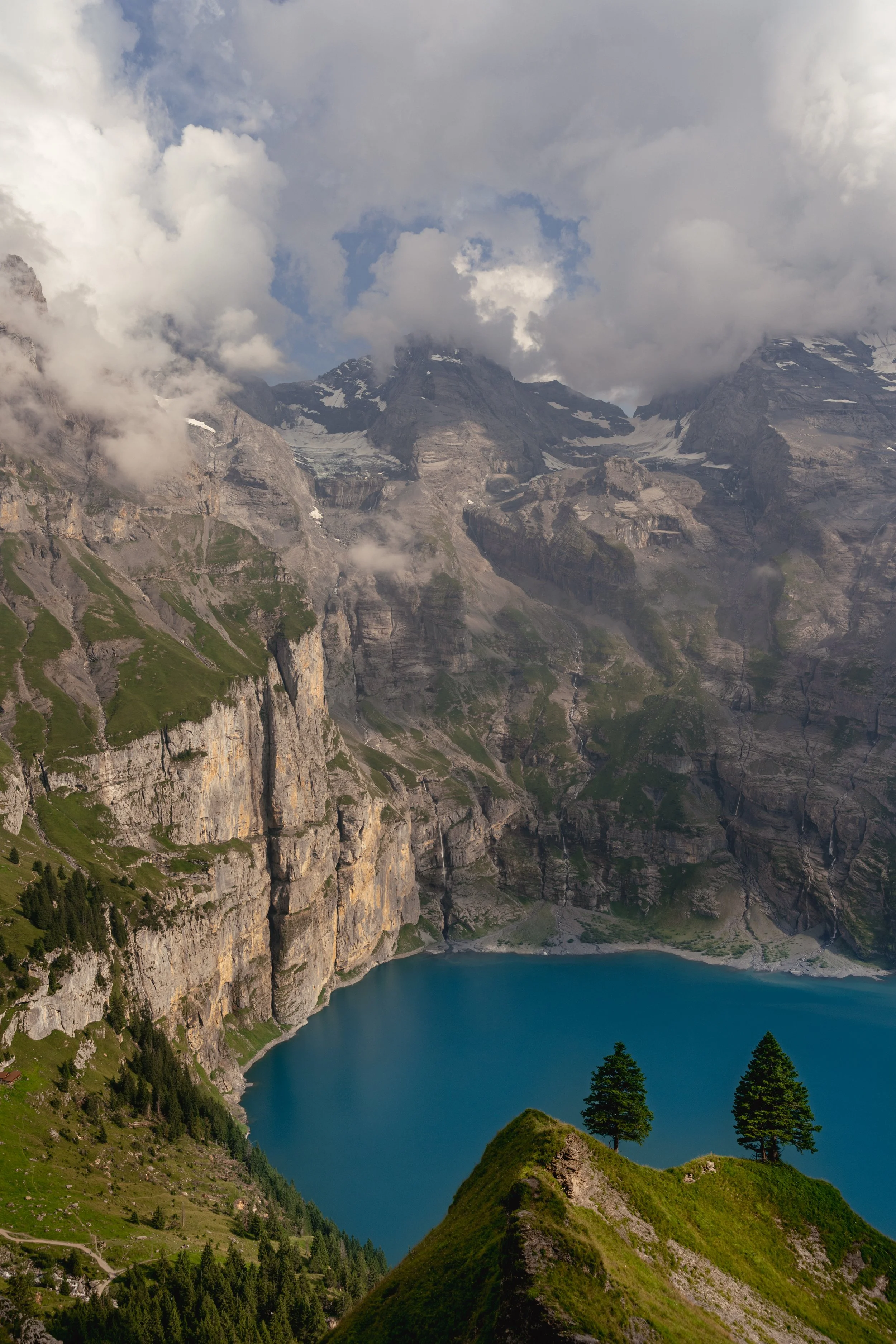 Berglandschaft mit schneebedeckten Gipfeln, Wolken, einem See und grünen Hügeln mit Bäumen.